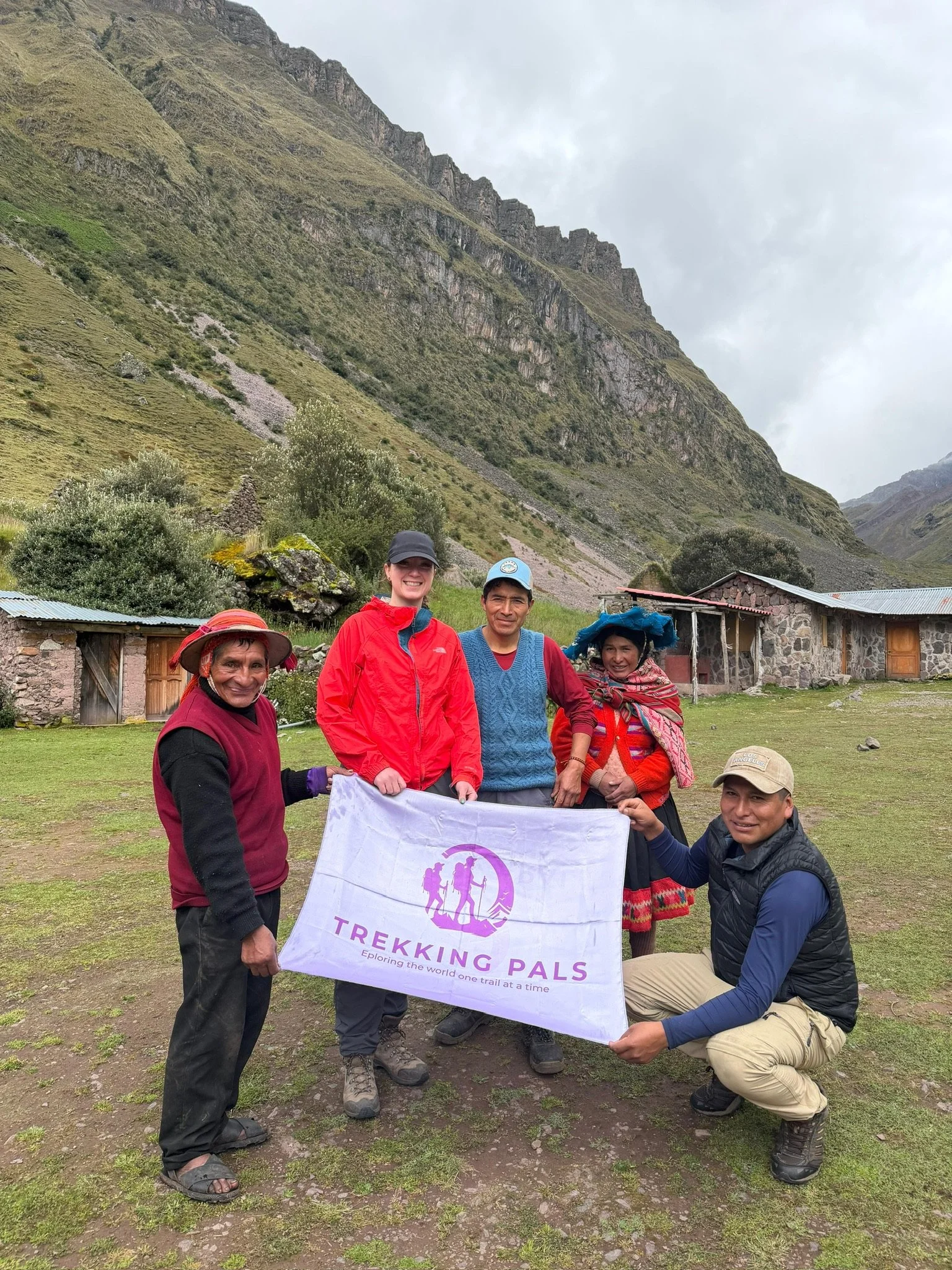 Hikers and local guides pose with a Trekking Pals banner beneath dramatic Andean mountains and stone buildings.