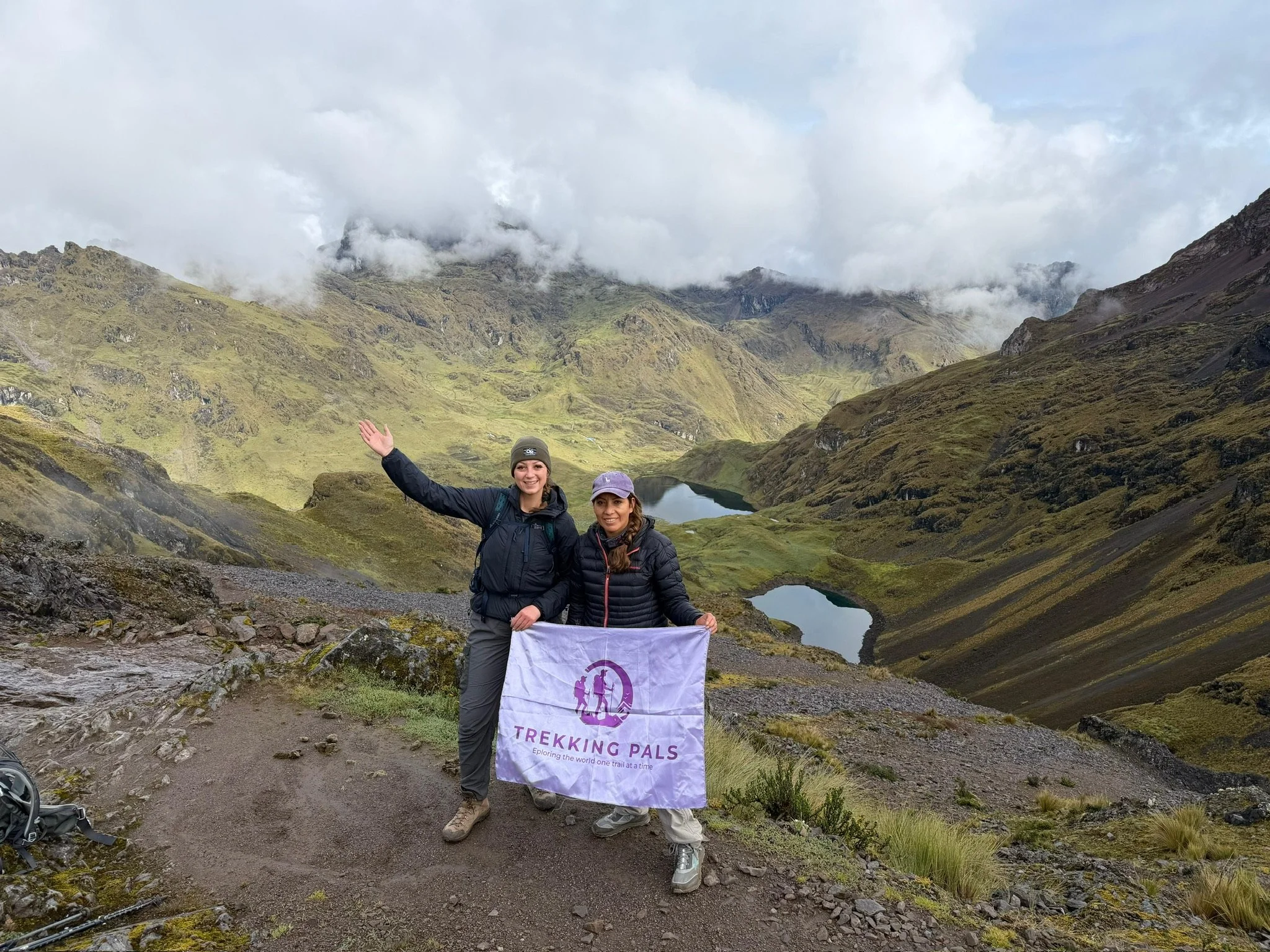Two hikers hold a Trekking Pals flag on a mountain trail overlooking two alpine lakes and cloud-covered peaks.
