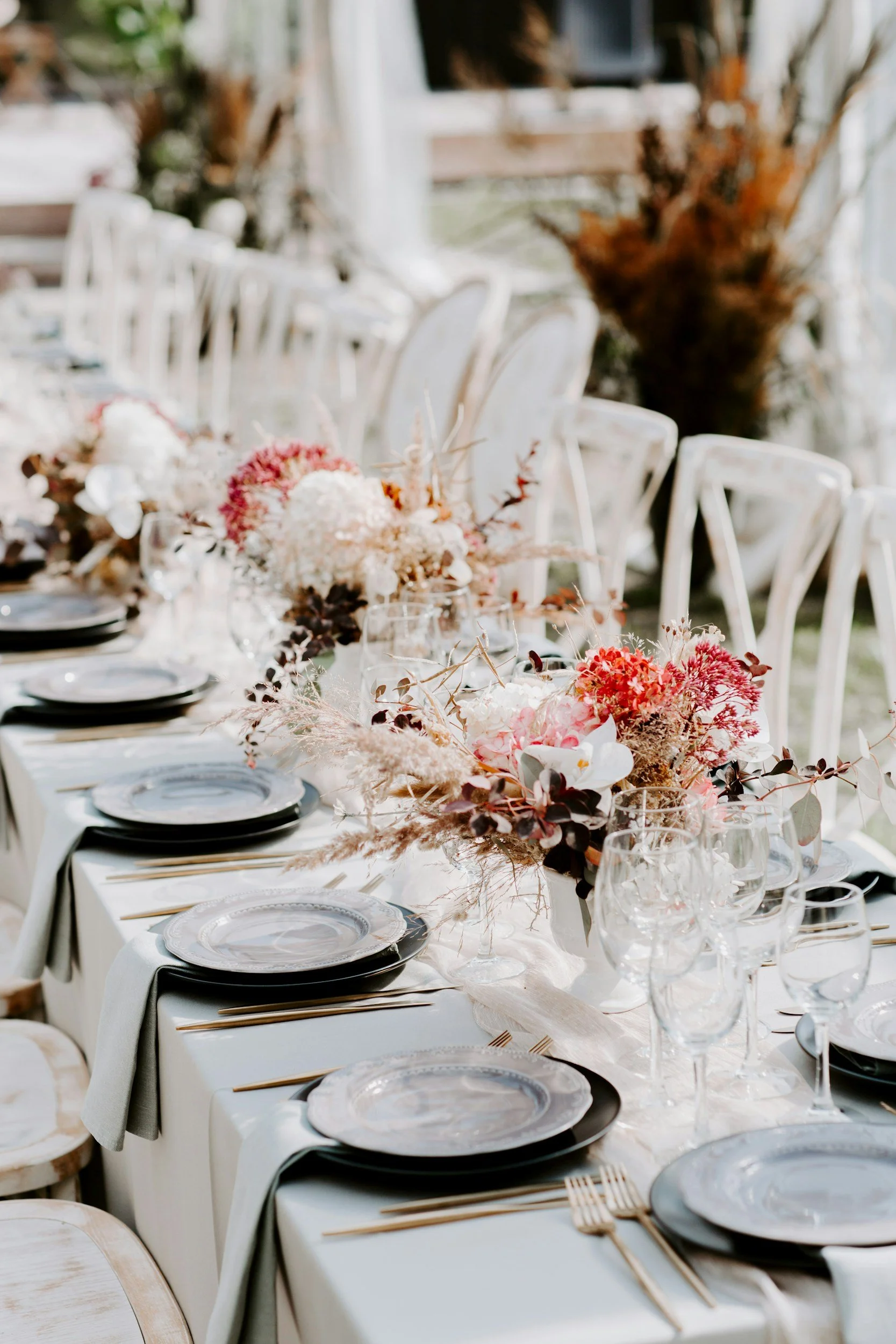 Sacramento Wedding with pink, white, and burgundy floral arrangements, set with black and clear glass plates, gold cutlery, and various wine glasses, with white chairs and soft natural lighting.
