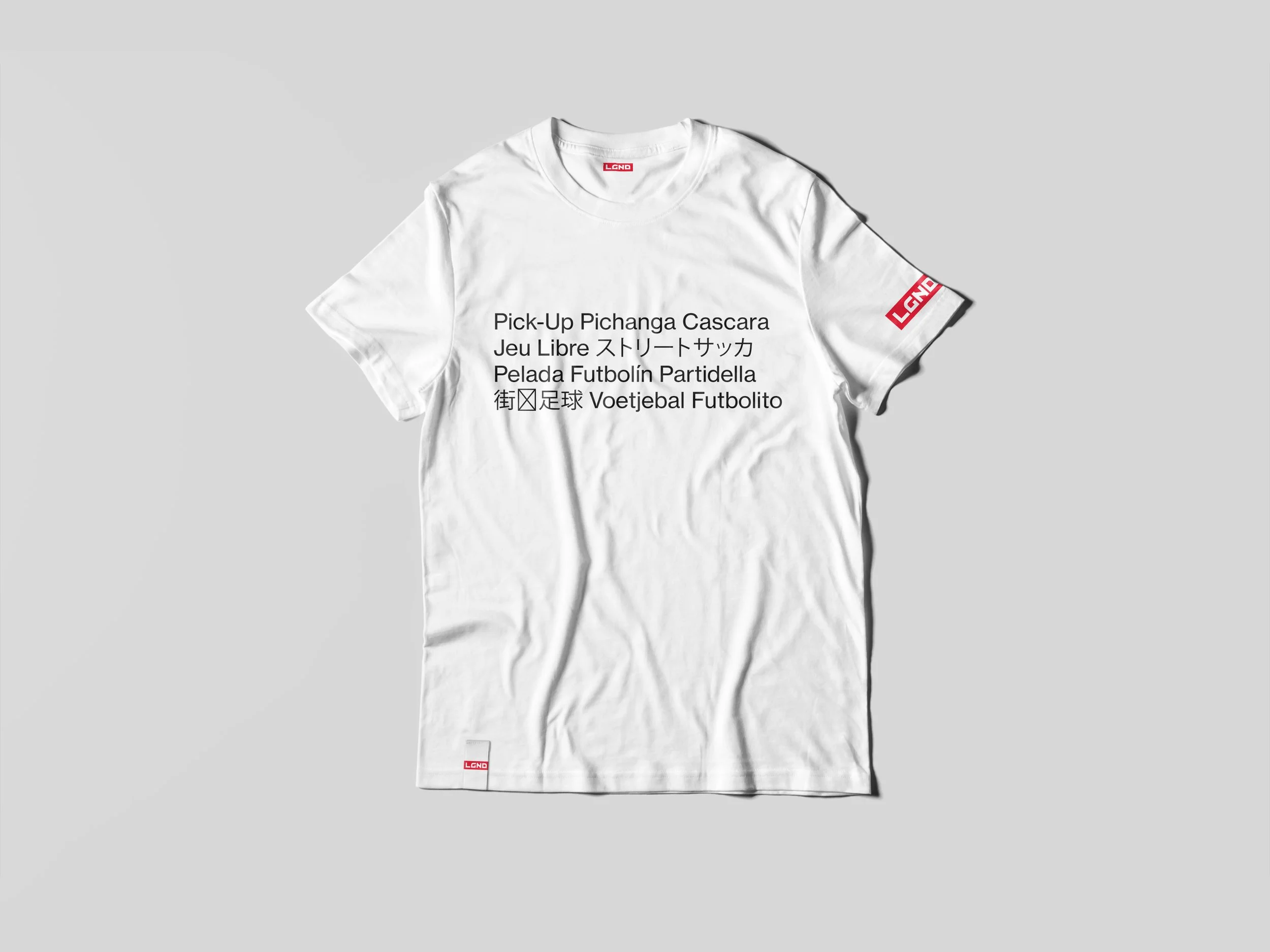 White T-shirt with black text in multiple languages and red logos, laid flat on a light background.