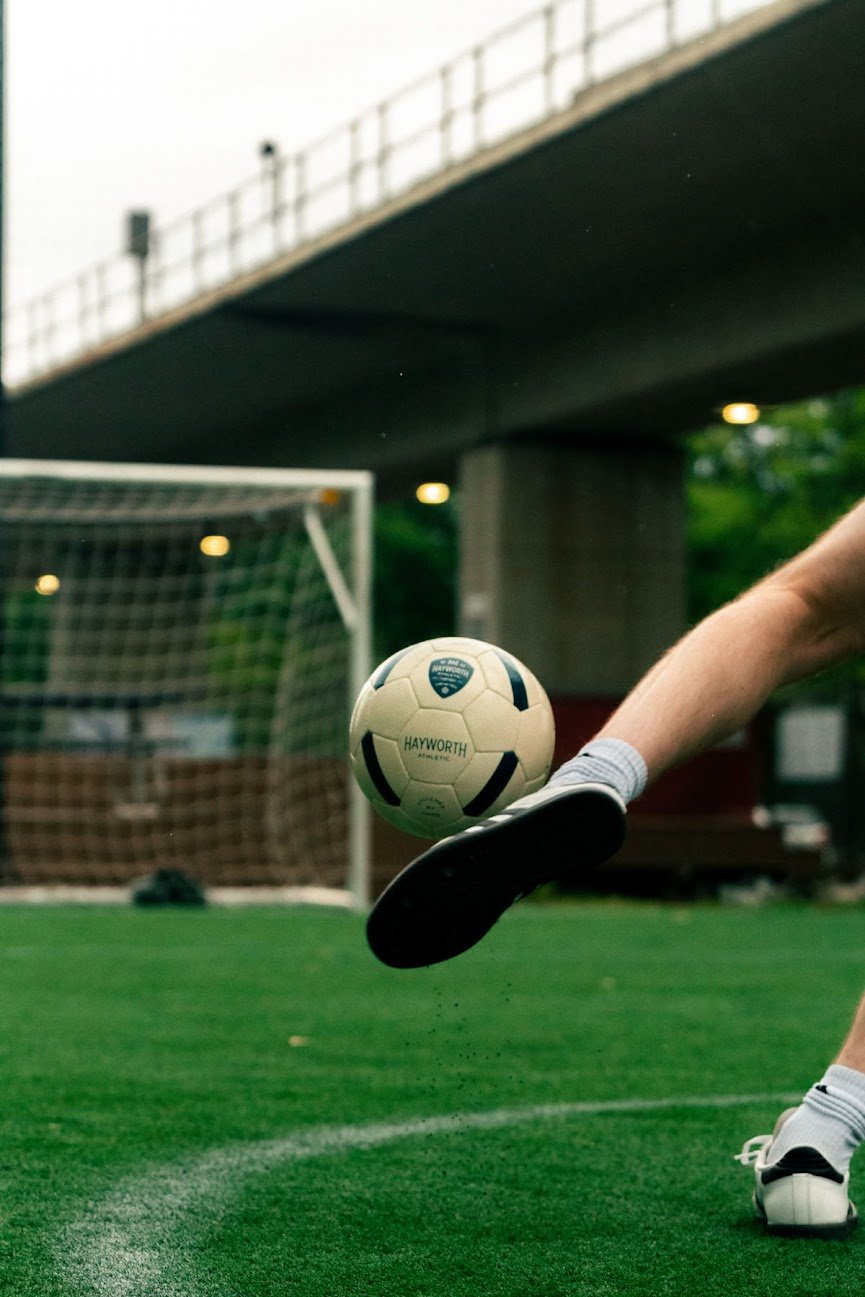 Close-up of a soccer ball kicking towards the goal on a green field, with a person wearing white shoes and athletic socks under a bridge in the background.