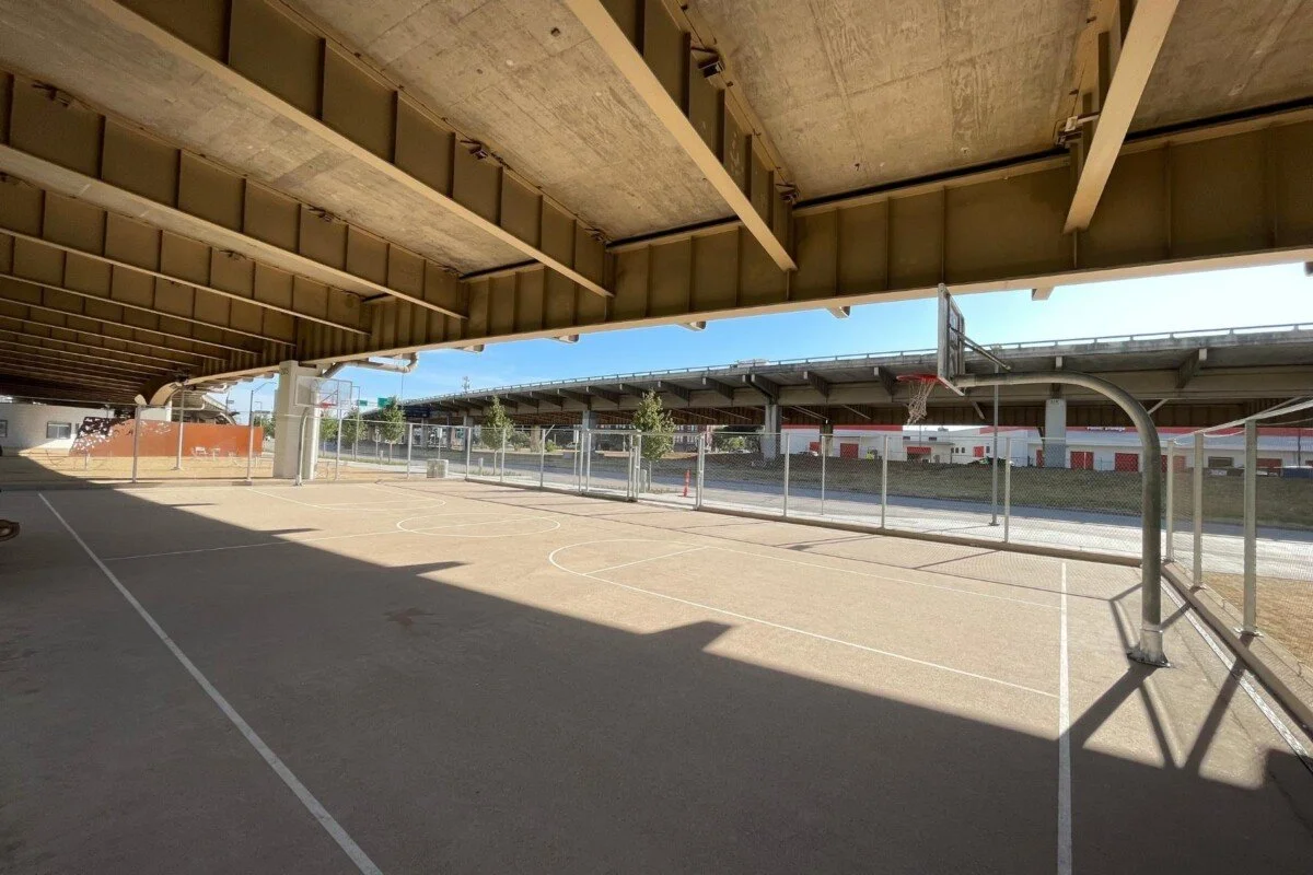 Underneath a highway overpass, an outdoor basketball court with a chain-link fence, basketball hoops, and white court lines. Bright sunlight casts shadows across the court.