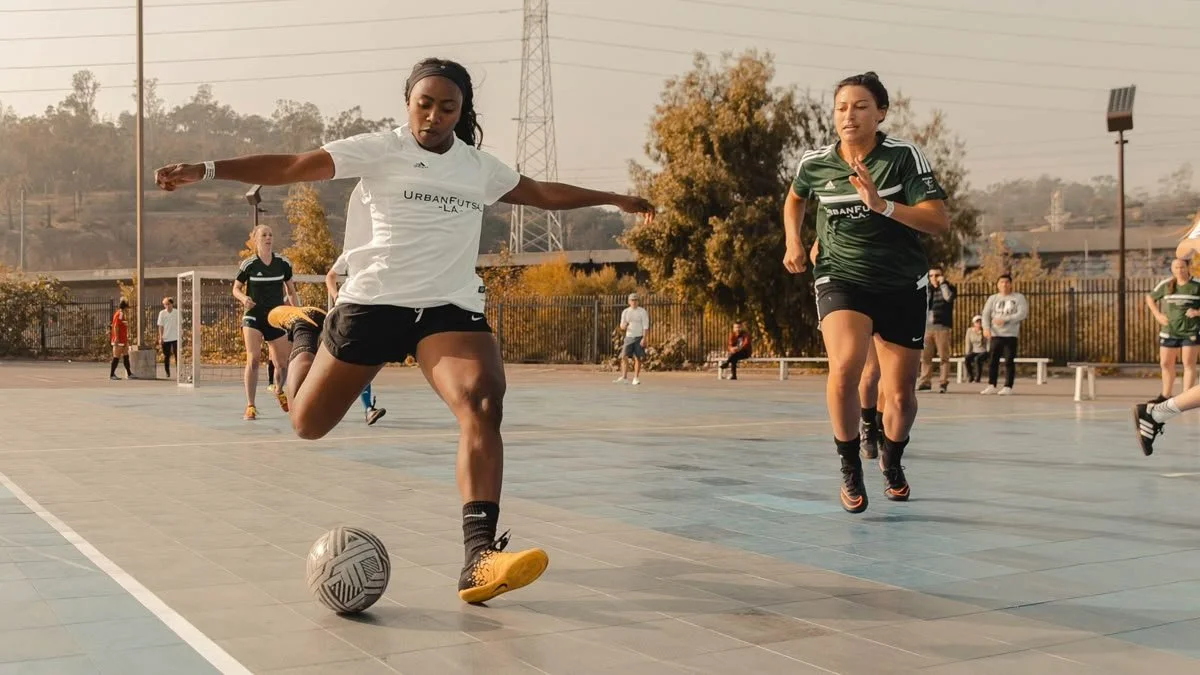 Two women playing soccer on an outdoor court, one in a white jersey kicking the ball and the other in a green jersey running.