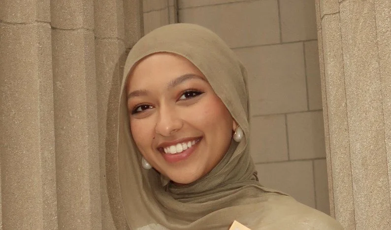 A woman wearing a beige hijab and pearl earrings, smiling, standing against a stone wall.