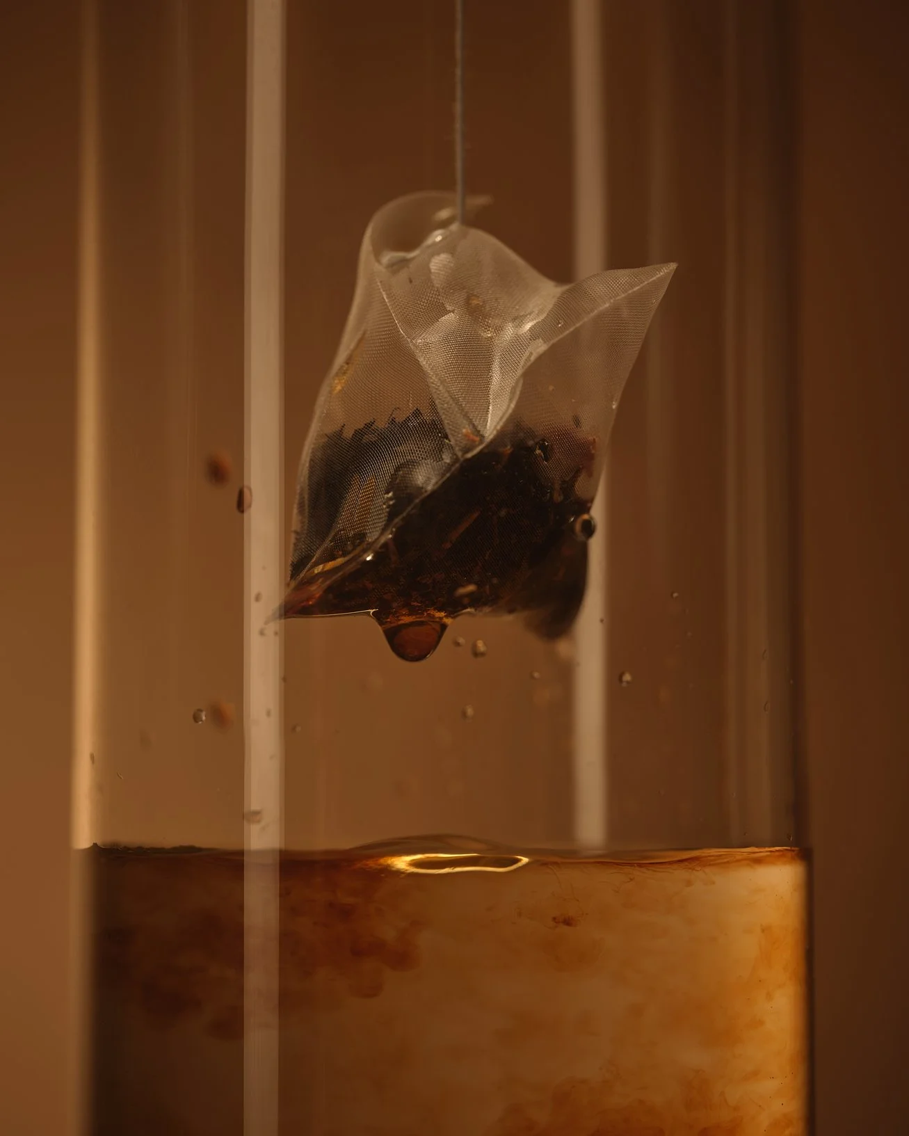 A tea infuser with tea leaves inside, hanging in a cup with hot water, with a warm brown background.