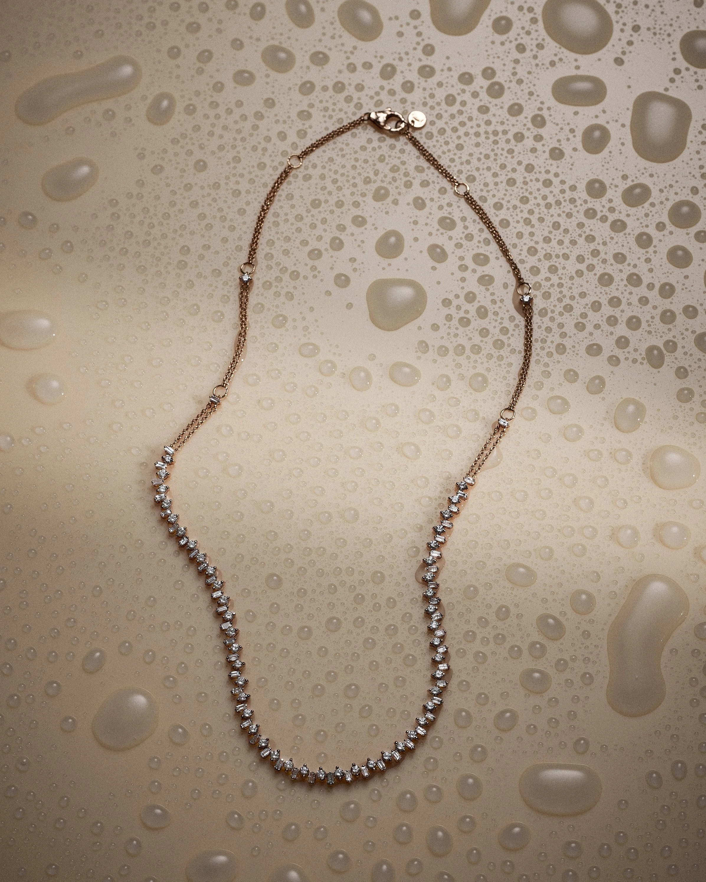 A diamond necklace with a delicate chain and a row of sparkling diamonds laid out on a beige surface with water droplets.