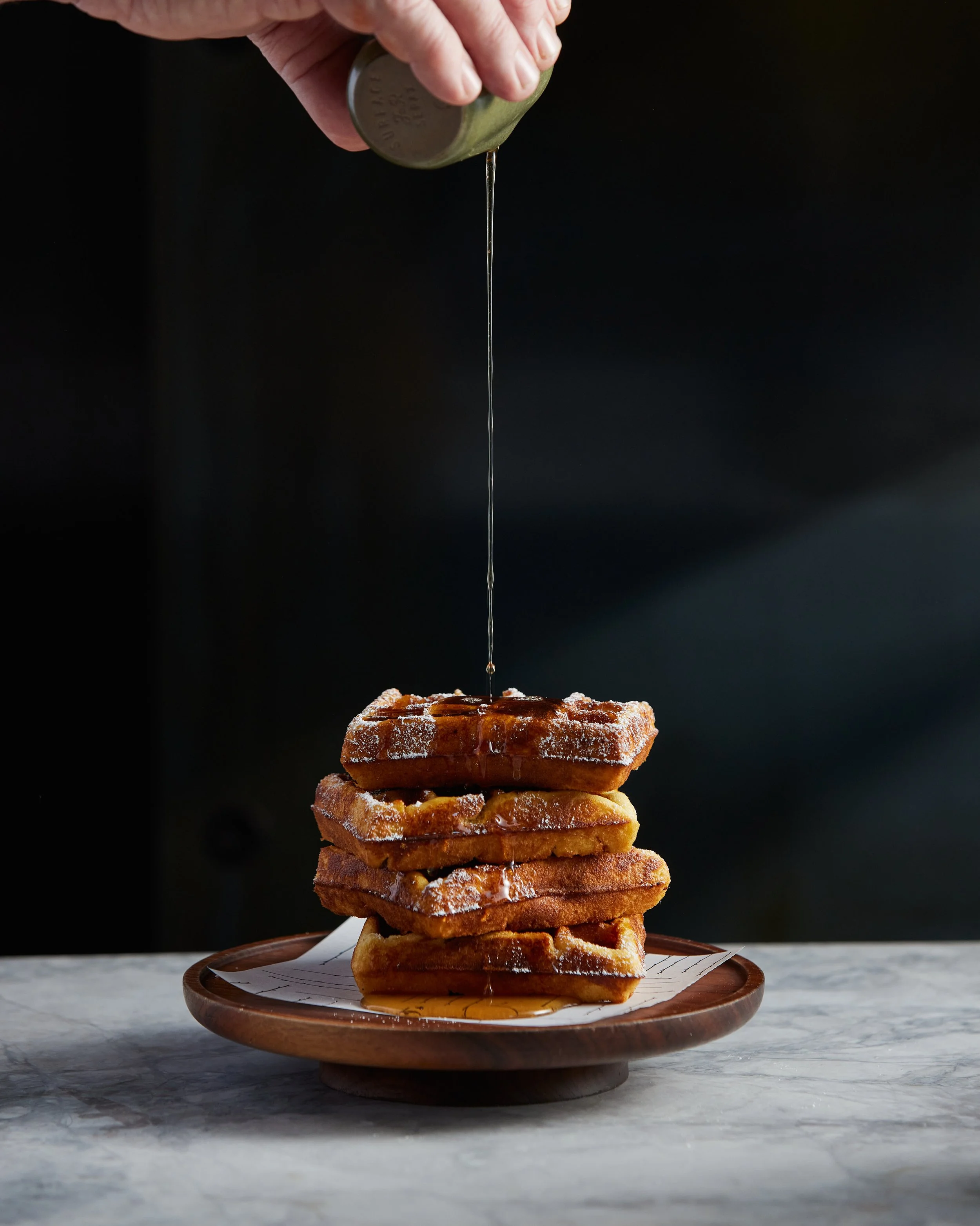 Stack of four waffles on a wooden plate, with syrup being drizzled from a metal container.