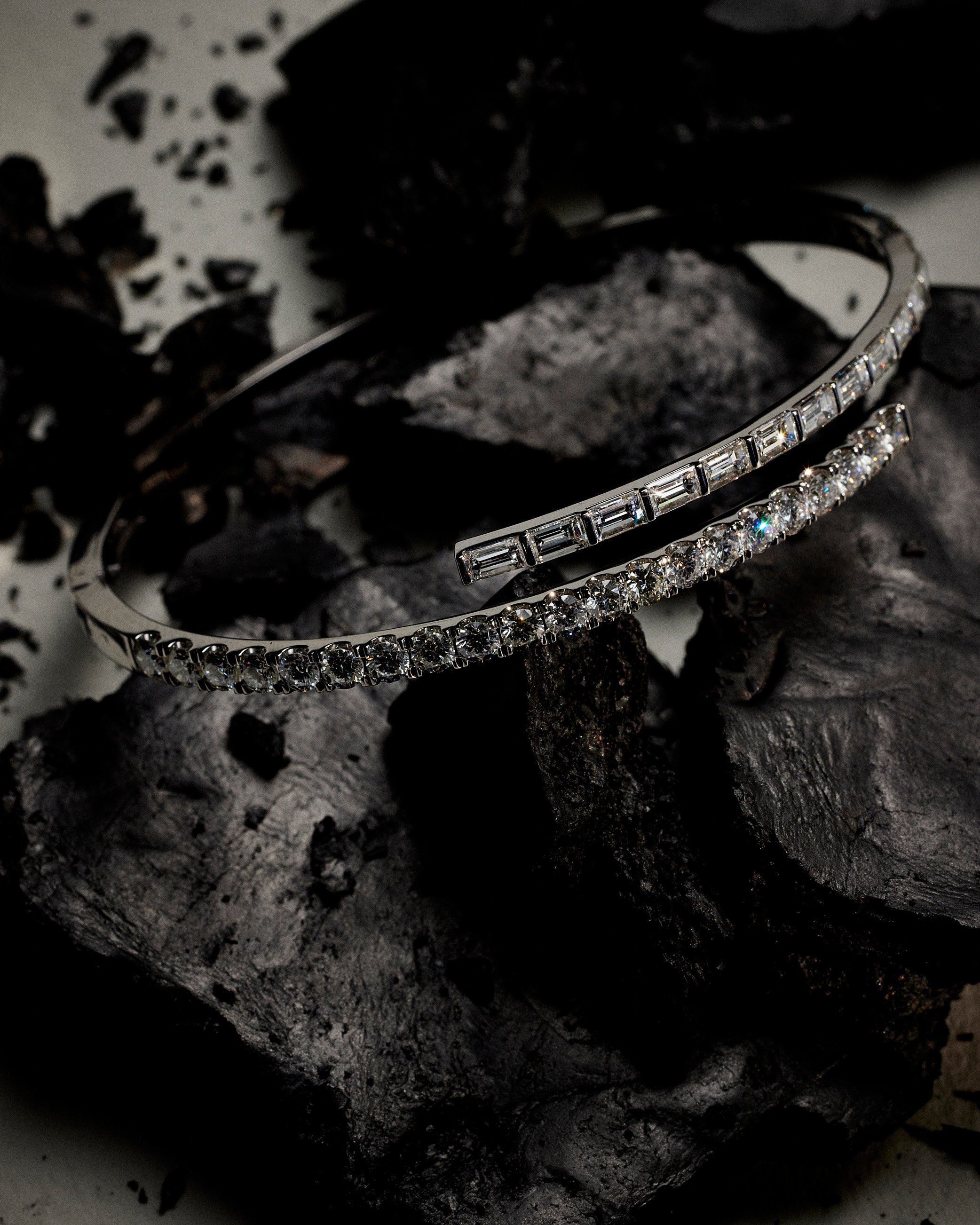 Two elegant silver bracelets with sparkling clear stones, resting on a textured black rock surface.