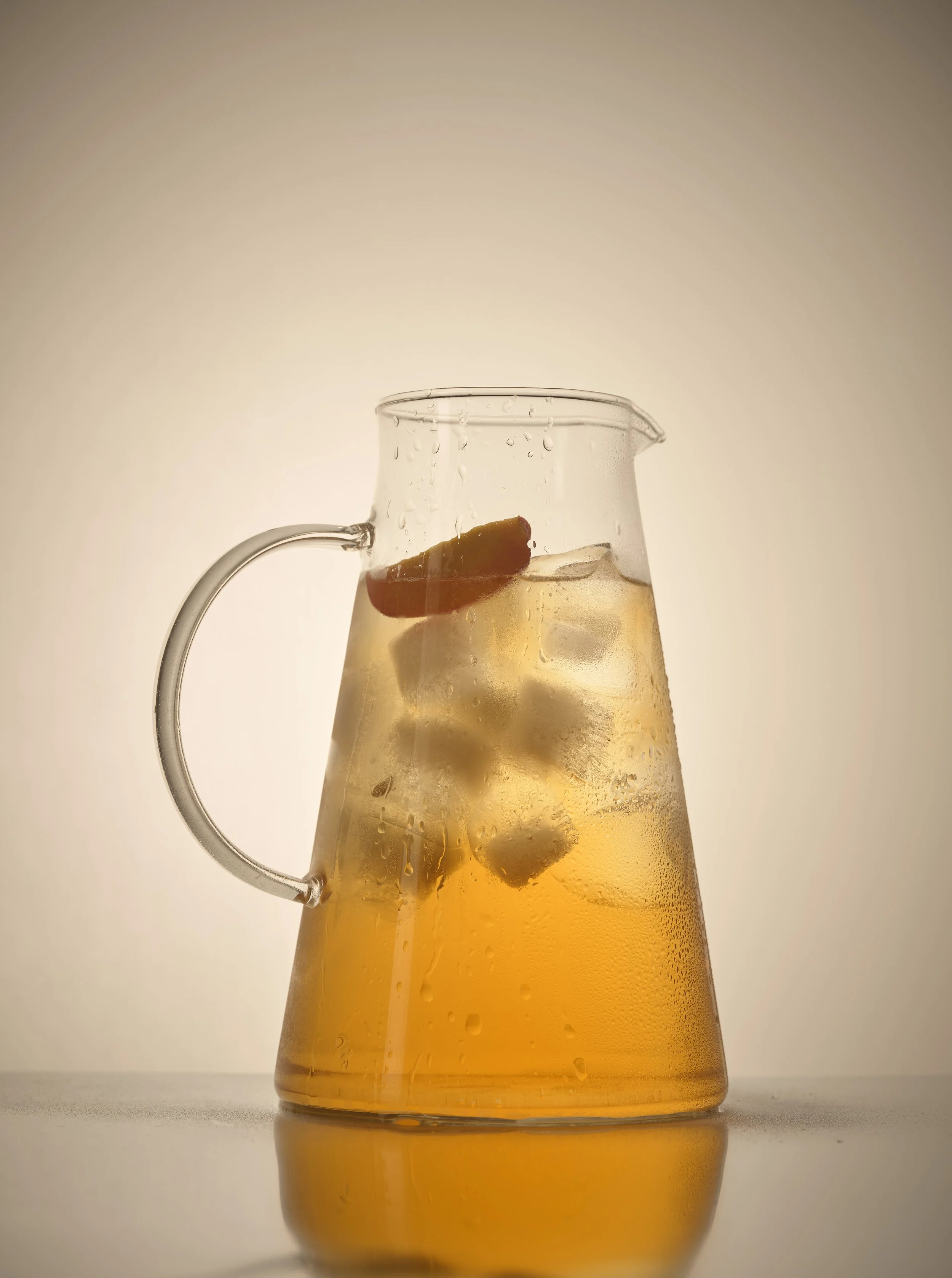 A glass pitcher filled with a cold, light amber-colored beverage with ice cubes and a lemon slice inside, condensation on the outside.