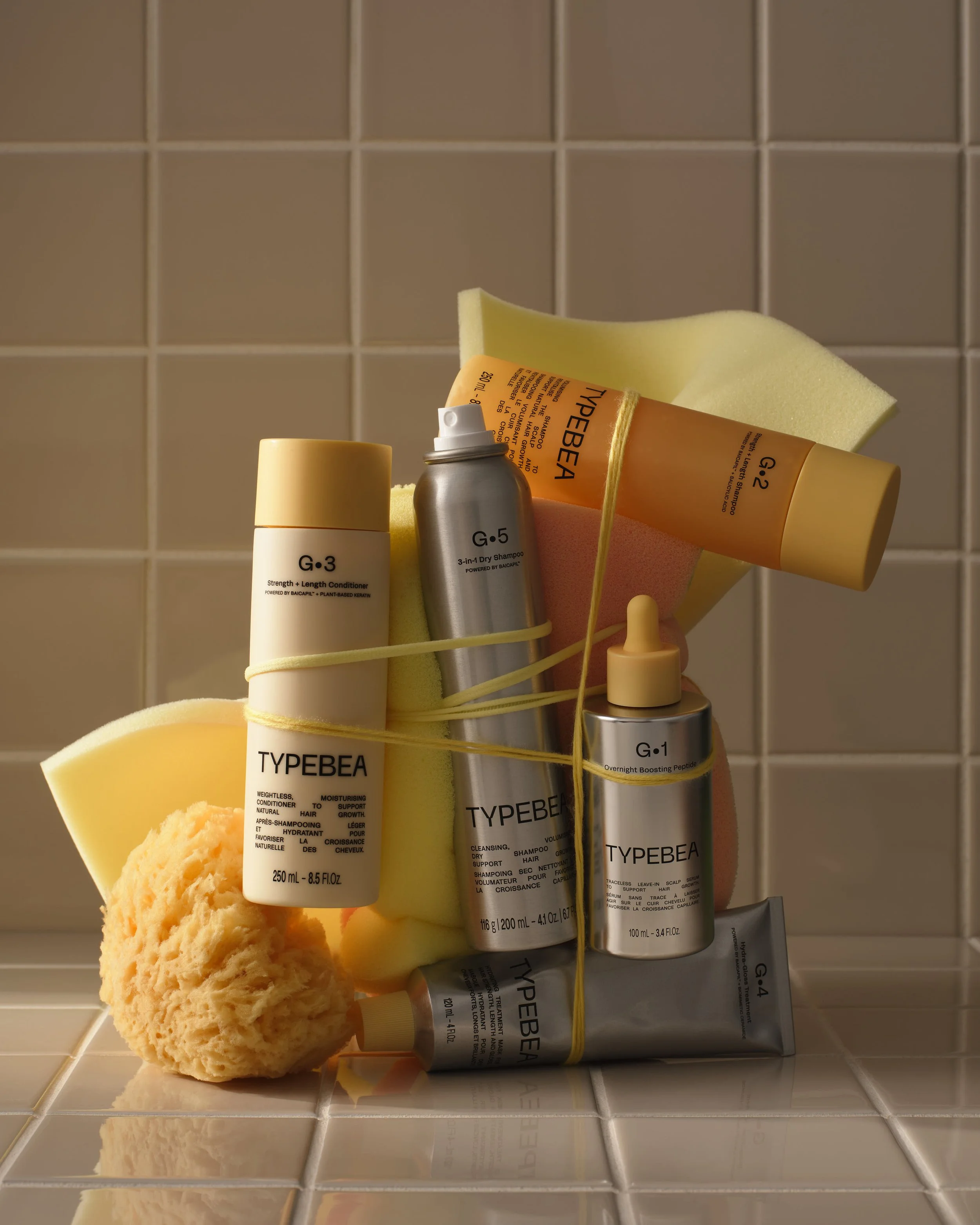A collection of hair care and styling products, including bottles of shampoo, conditioner, and styling sprays, along with a yellow sponge and a natural sea sponge, all arranged on a tiled bathroom shelf.