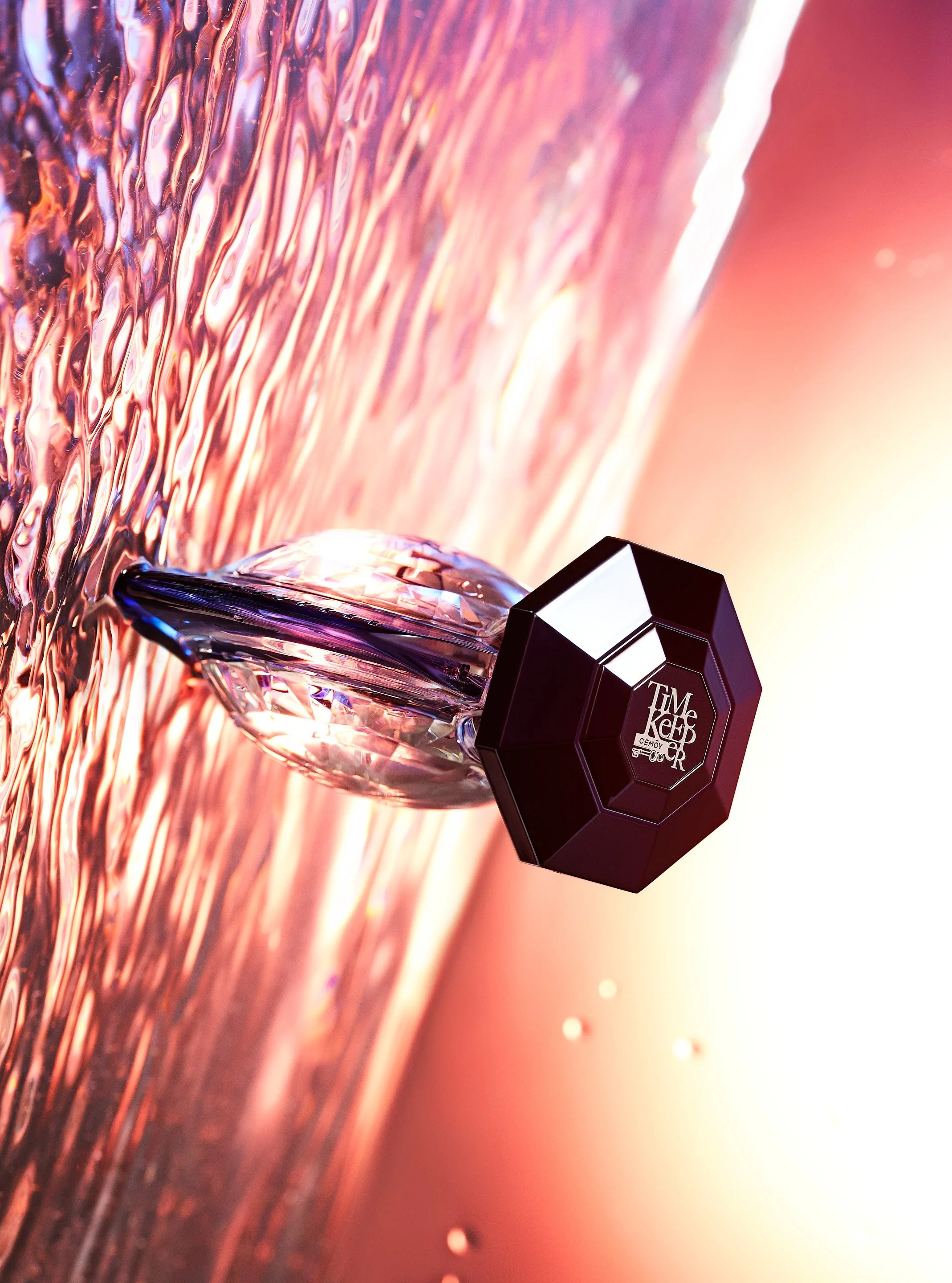 Close-up of a perfume bottle with a black, octagonal cap, resting on a textured, shimmering pink surface with light reflections.