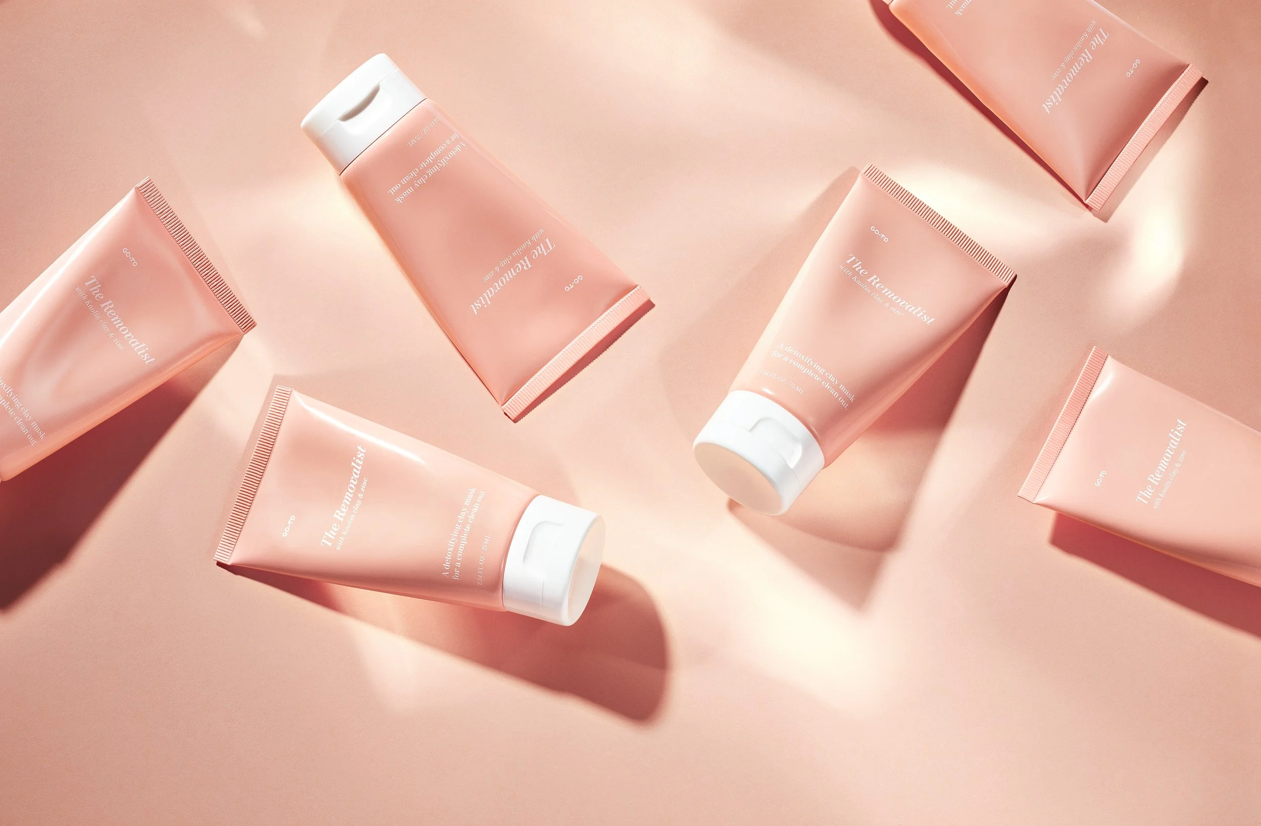 Several pink tubes of skincare cream labeled "The Removatalist" are scattered on a pink background with soft shadows.