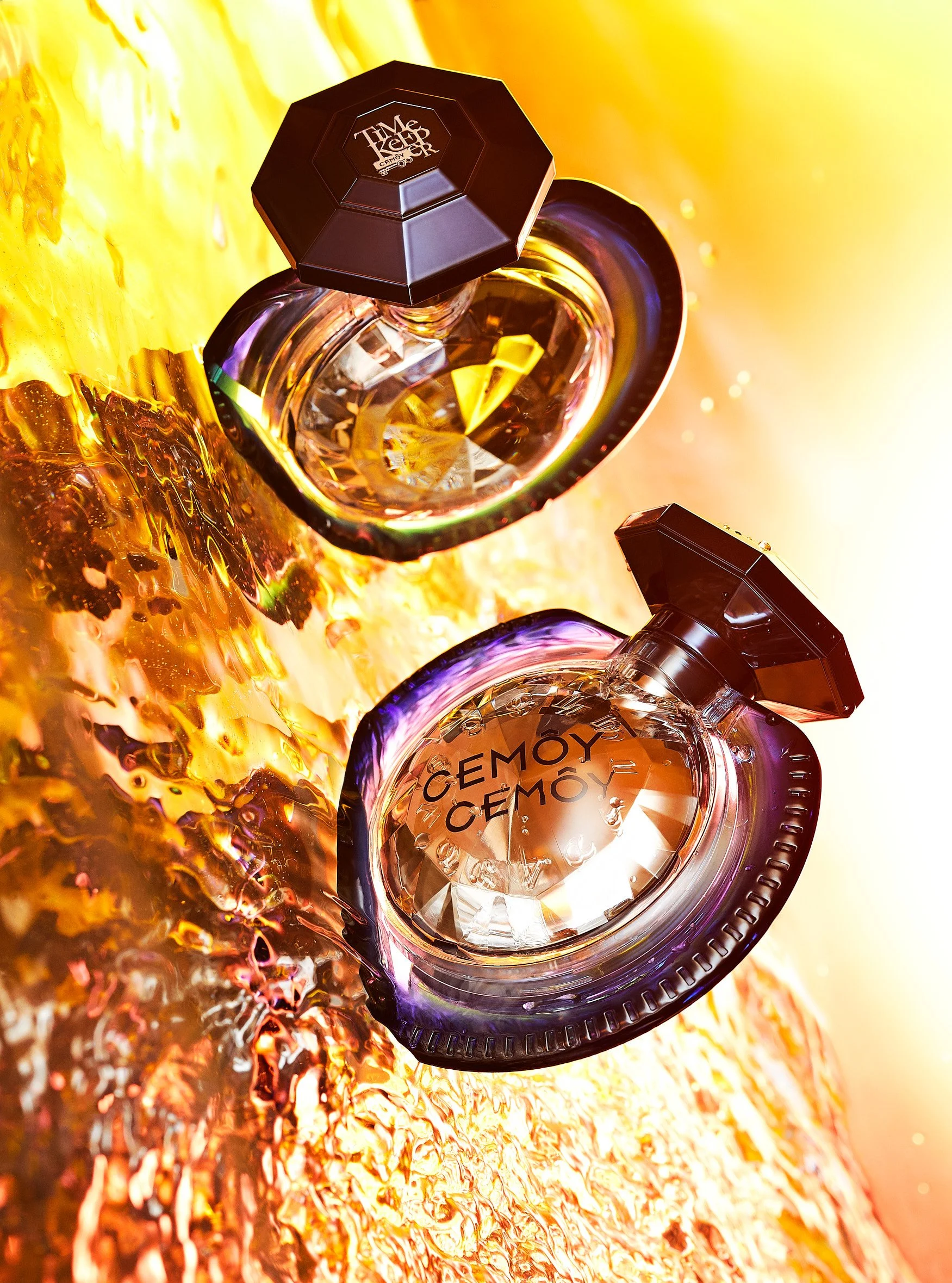 Two round glass containers with black lids and the word 'CÉMOY' printed on them, placed on a vibrant golden-yellow surface with water splashes and warm lighting creating a bright, energetic atmosphere.