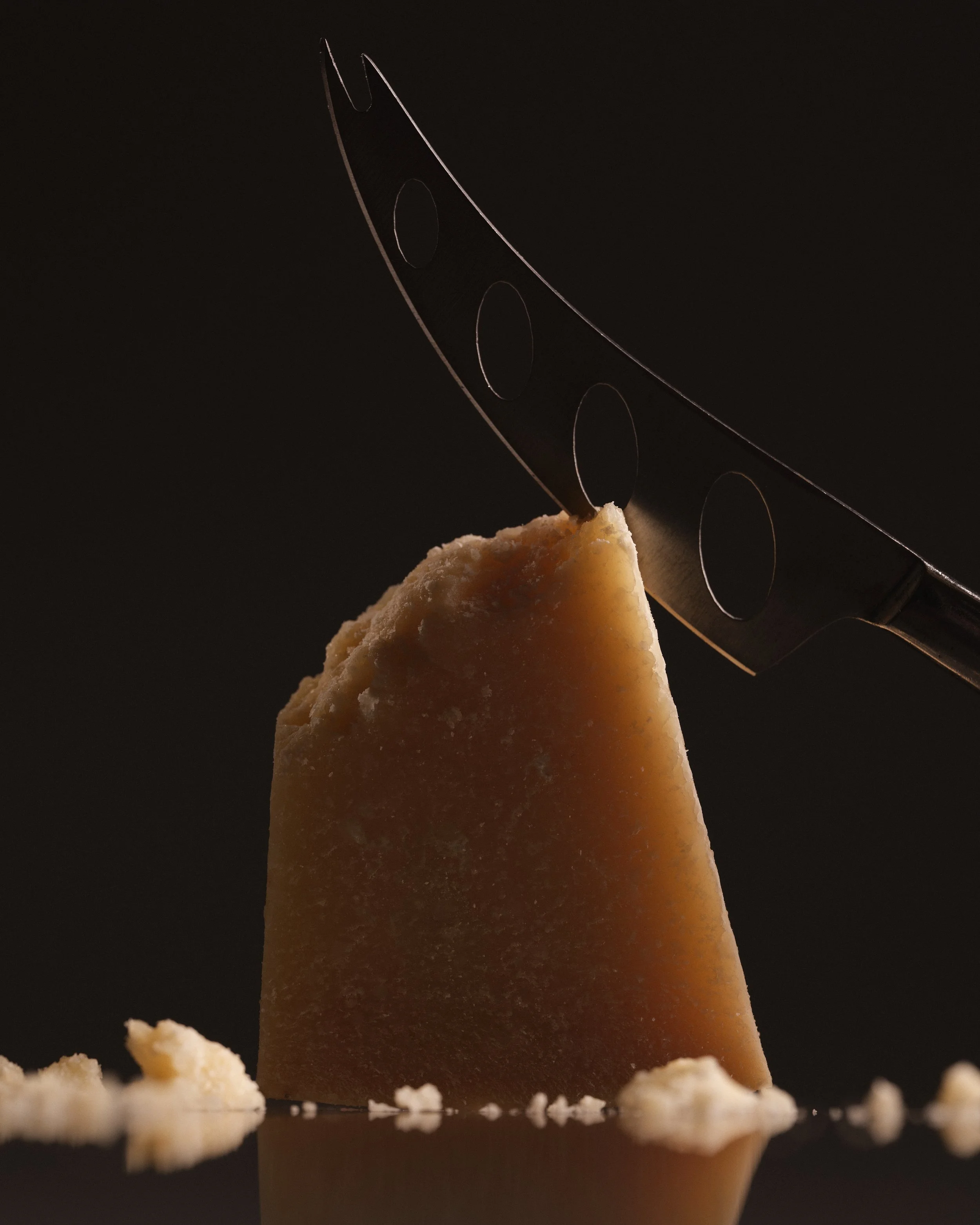 A chunk of cheese with a cheese slicer inserted into it, on a dark surface with some crumbs around.