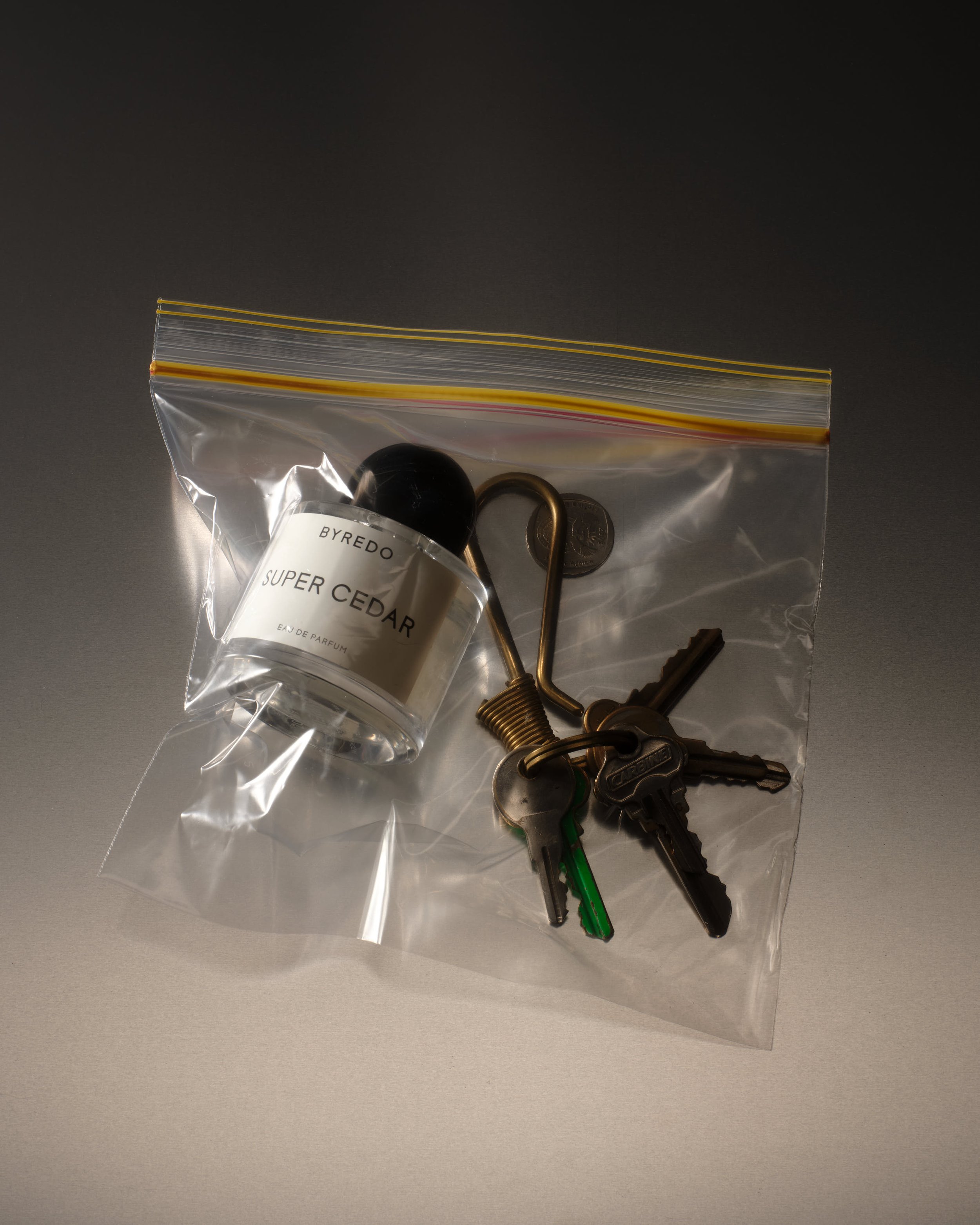 A clear plastic bag containing black and green keys, a small black bottle labeled 'BYREDO SUPER CEDAR', a gold-colored hook, and a coin, on a gray surface.