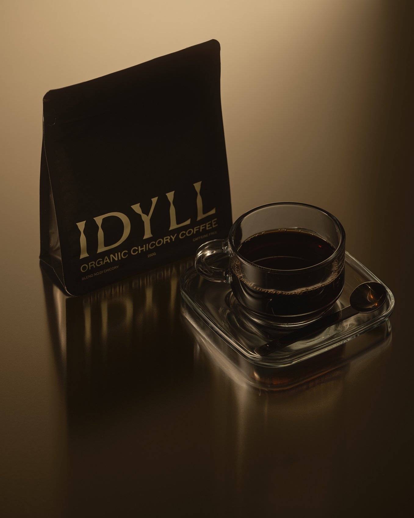 A bag of IDYLL organic chicory coffee, a clear glass cup filled with black coffee, and a small spoon on a glass tray, all placed on a metal surface with dim lighting.
