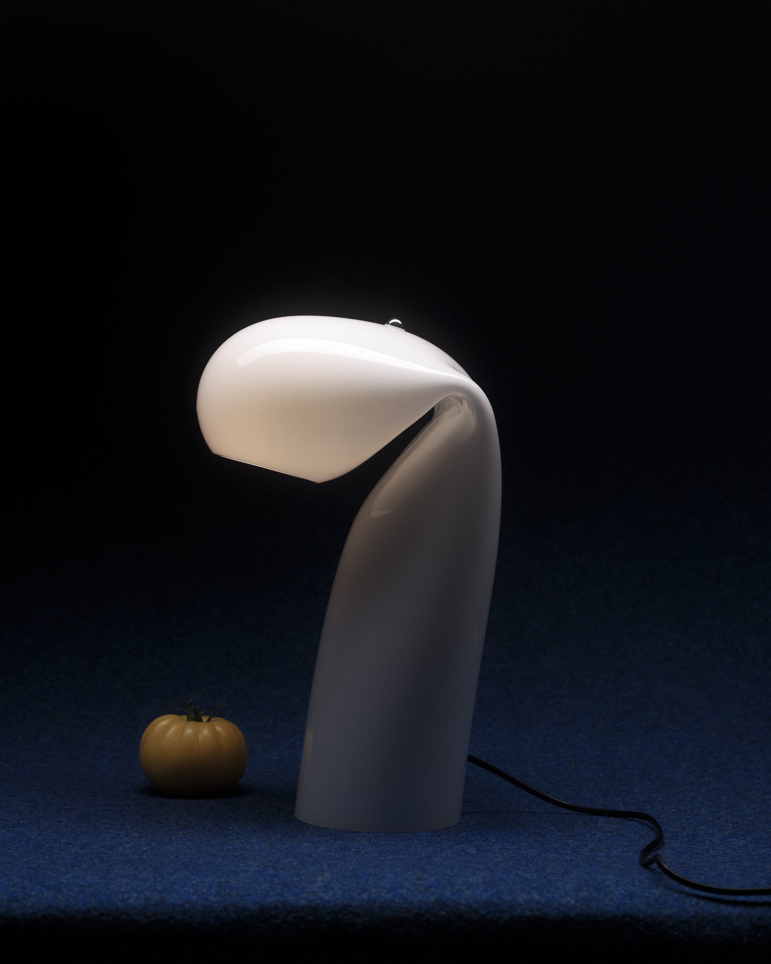 Modern white table lamp with curved design, positioned on a dark surface with a small yellow tomato nearby, against a black background.