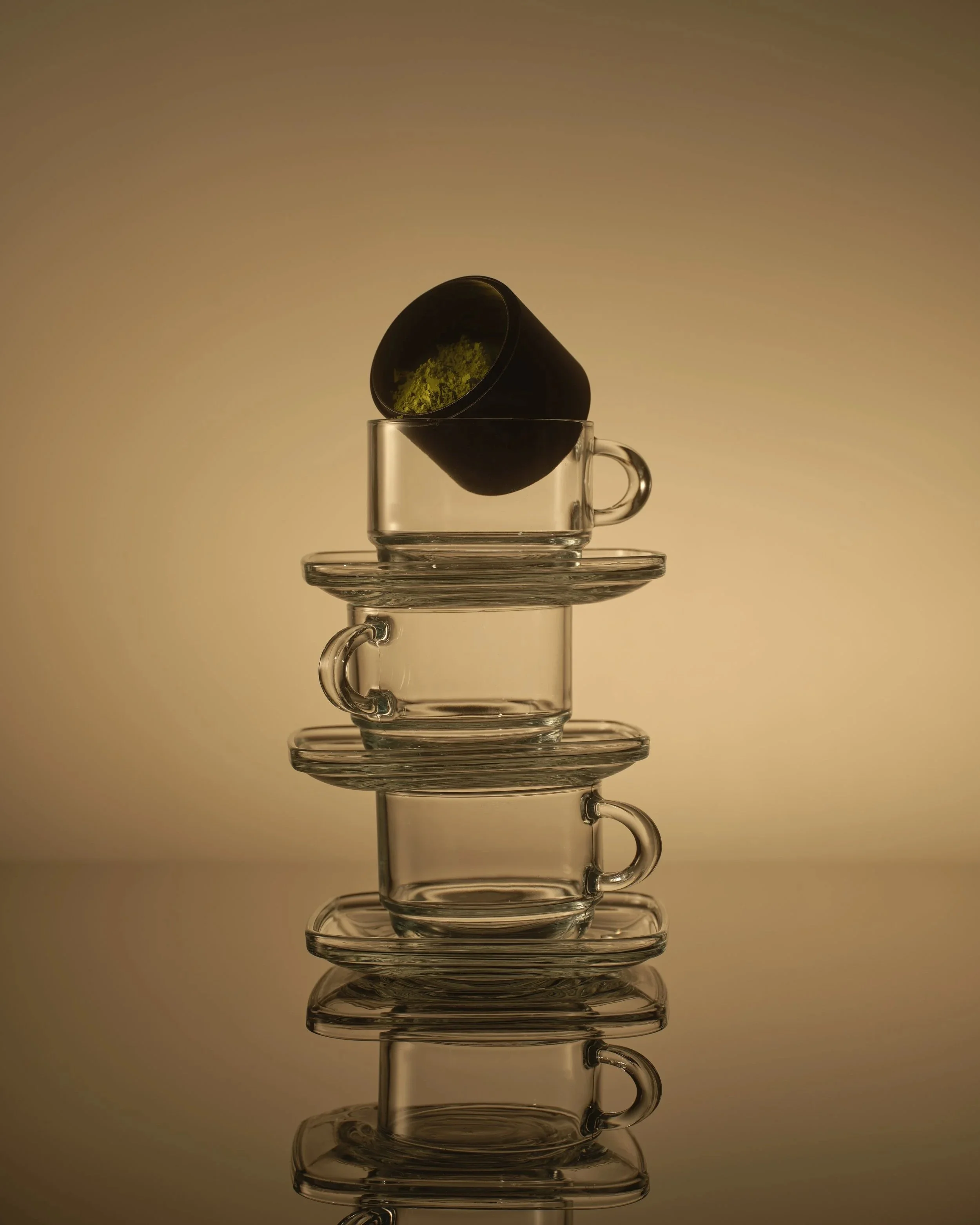 Stacked clear glass cups with a black cup on top containing green matcha powder, arranged in a vertical line against a plain background.