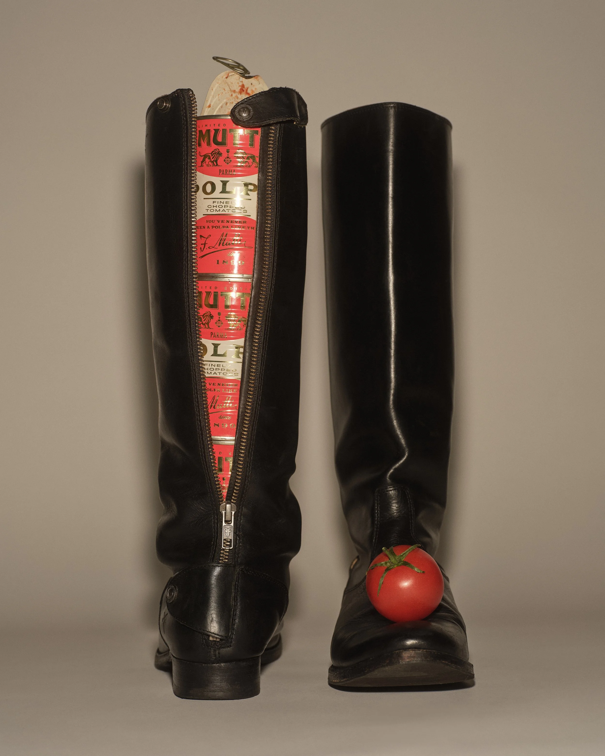 A pair of black leather boots with zipped zippers, one of which is partially open revealing a tomato and a block of cheese inside, with a tomato placed at the toe of one boot.