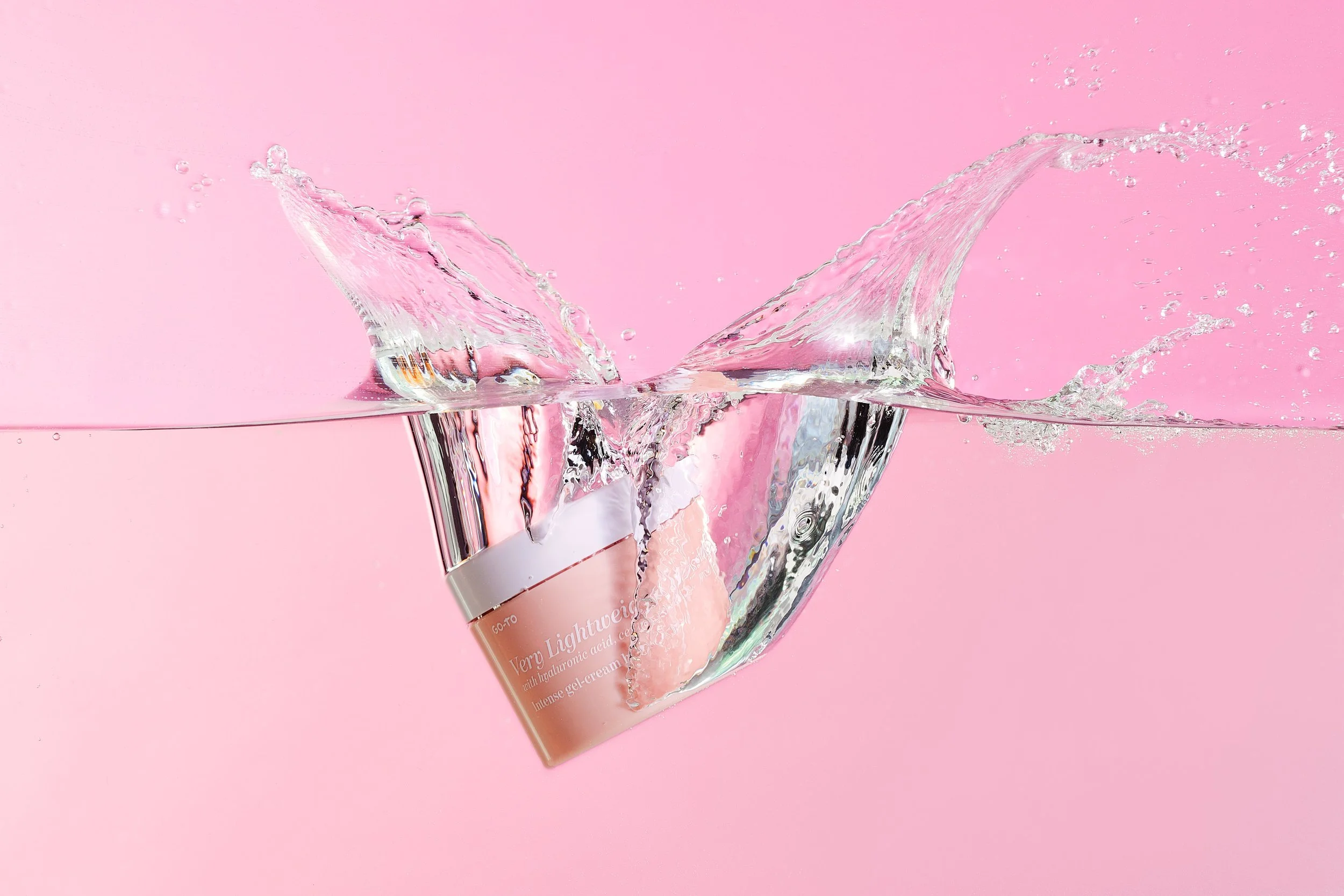A jar of Very Light Reveur gel cream sinking into water, creating a splash, against a pink background.
