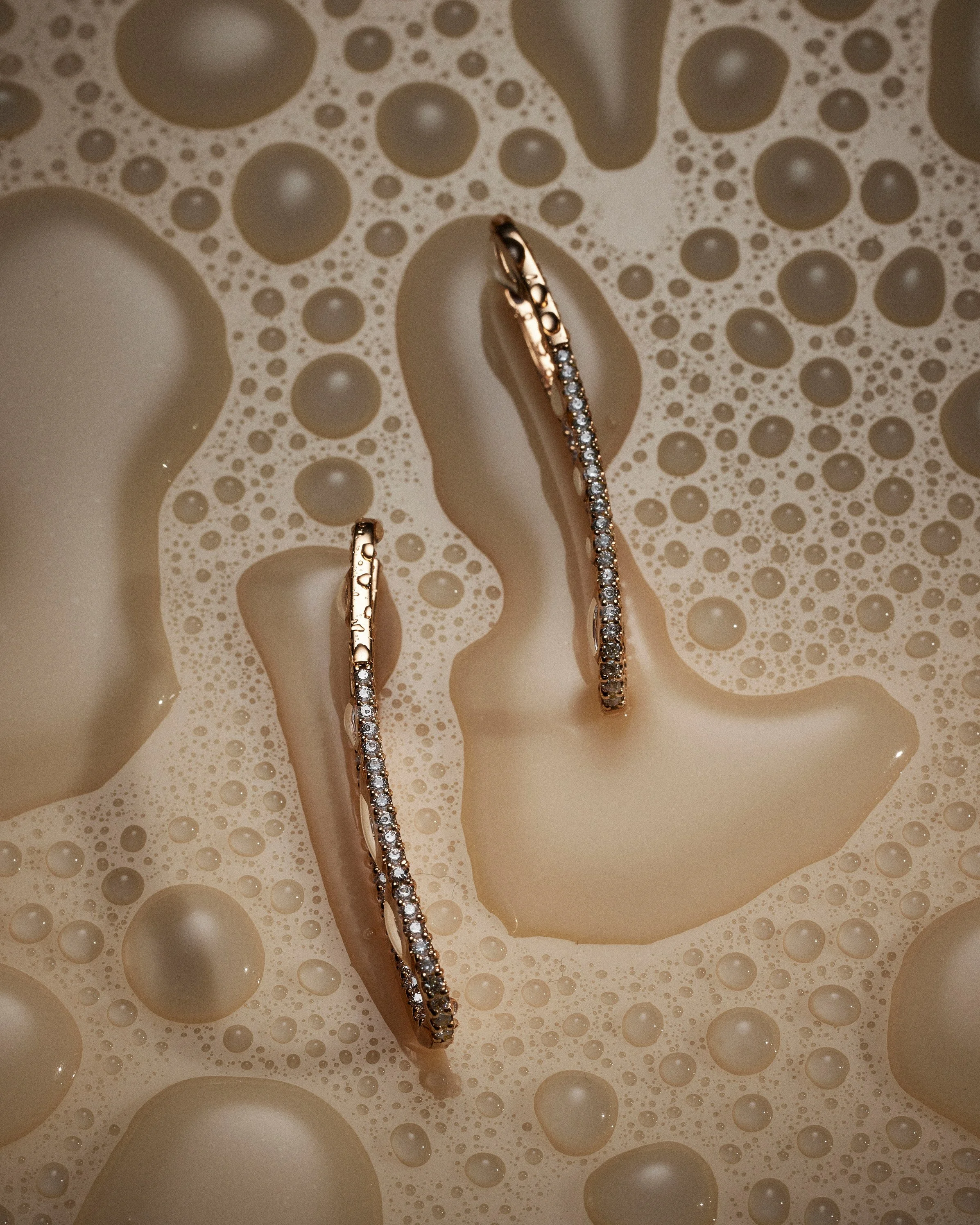 Gold hoop earrings with small diamonds resting on a surface of soapy bubbles.