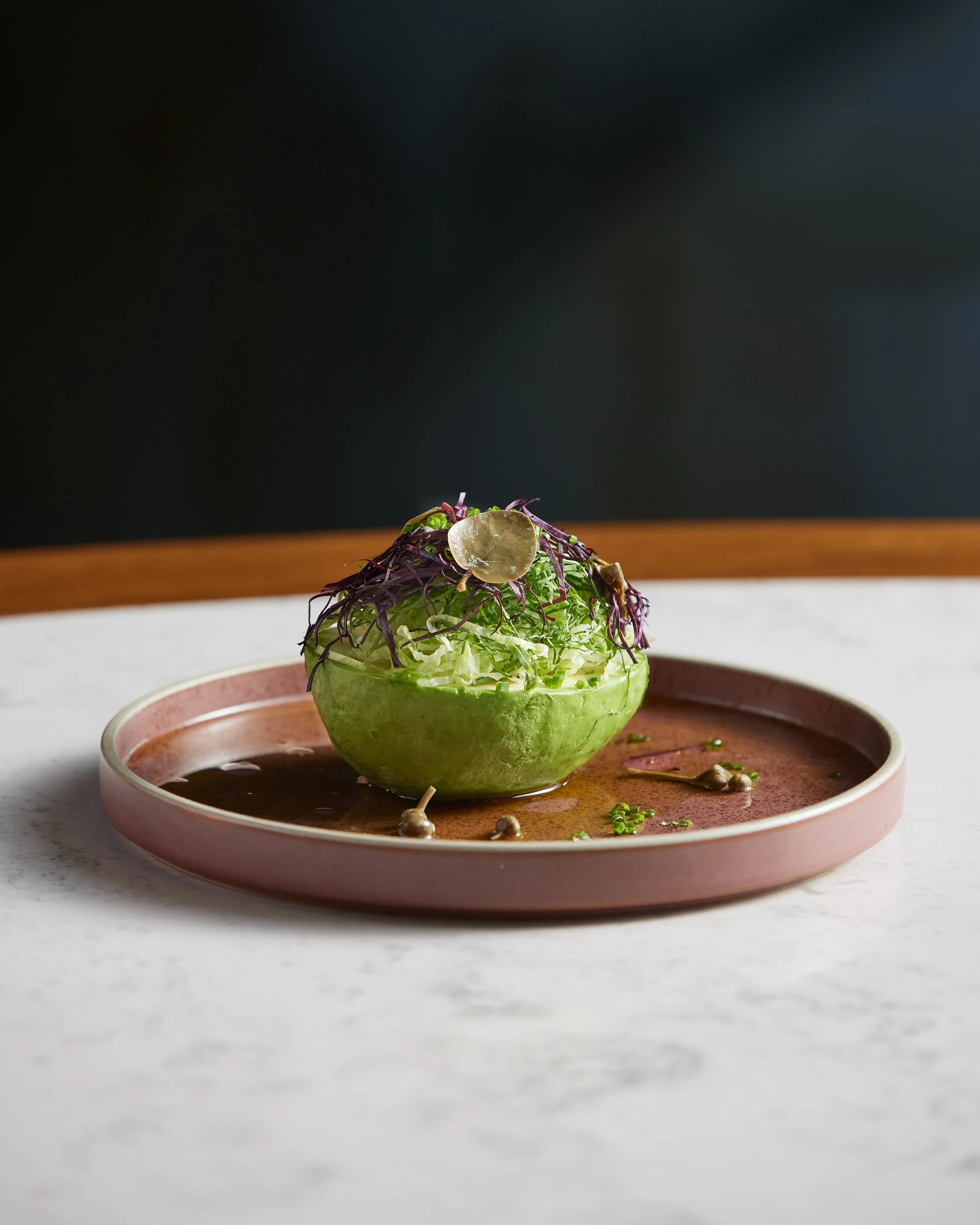 A bowl with a hollowed-out green apple filled with shredded lettuce, topped with purple shredded vegetables and a pickle slice, placed on a pink plate with a brown sauce and garnished with capers and herbs.