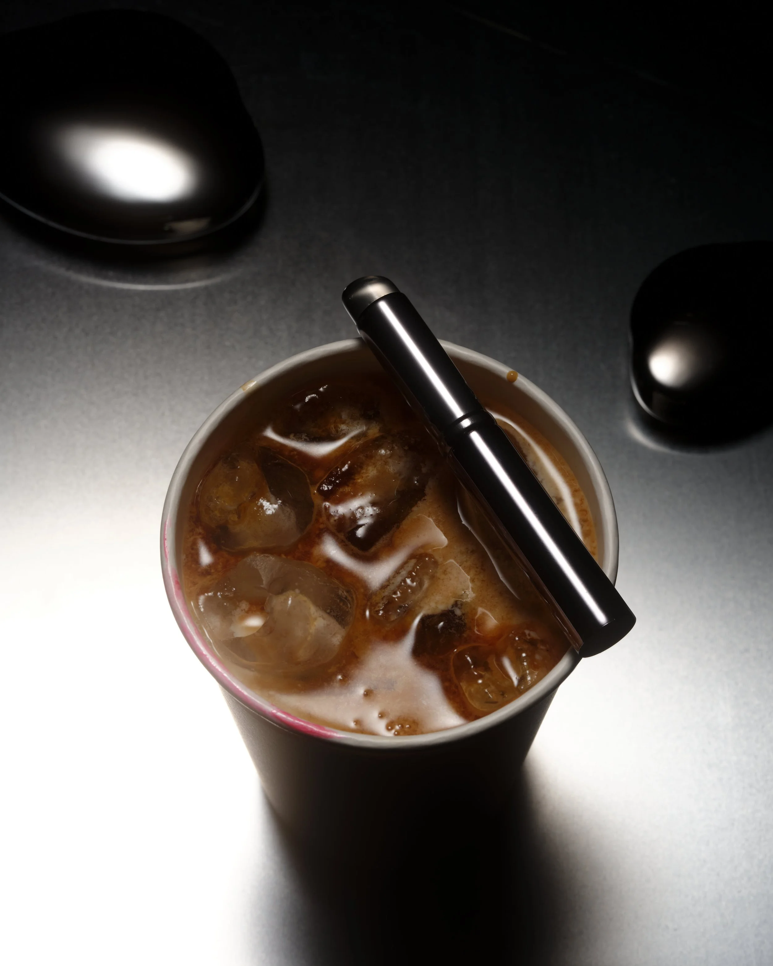 A cup of iced coffee with a black straw on a dark surface, with two black speakers in the background.