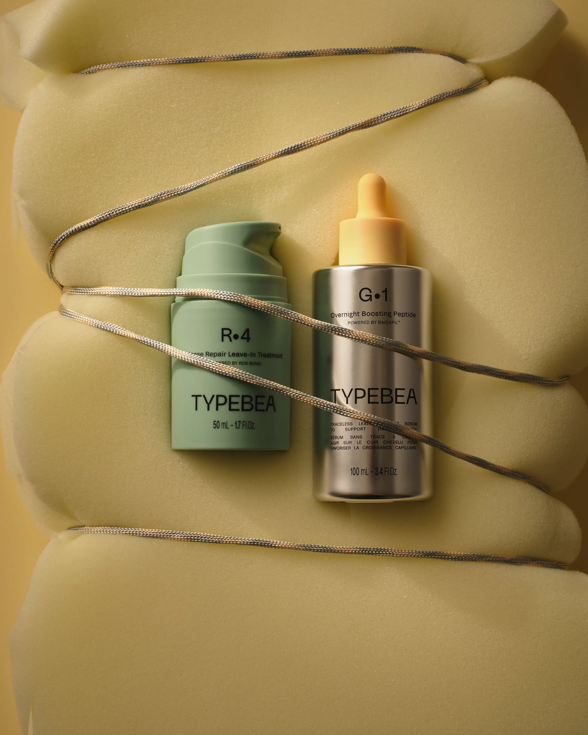 Two skincare bottles with pumps, one labeled 'R•4' and the other labeled 'G•1', placed on yellow foam with a silver cord draped over the foam.