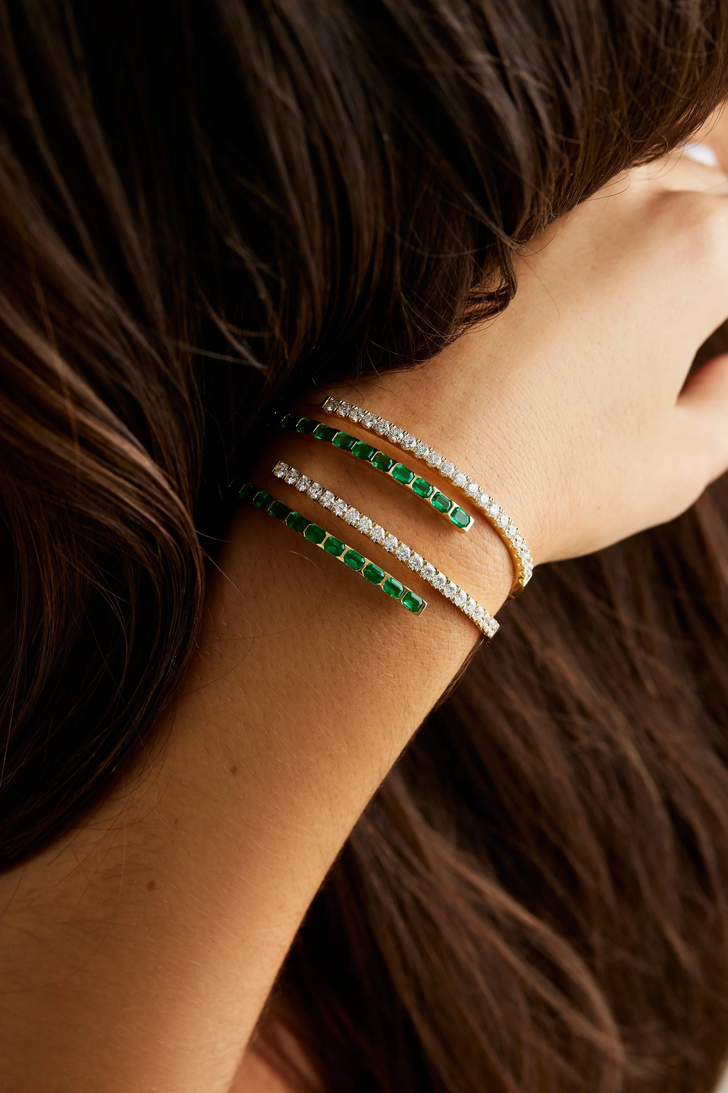 A woman's neck and face showing four layered bracelets: two with green gemstones and two with clear gemstones, against a backdrop of long brown hair.
