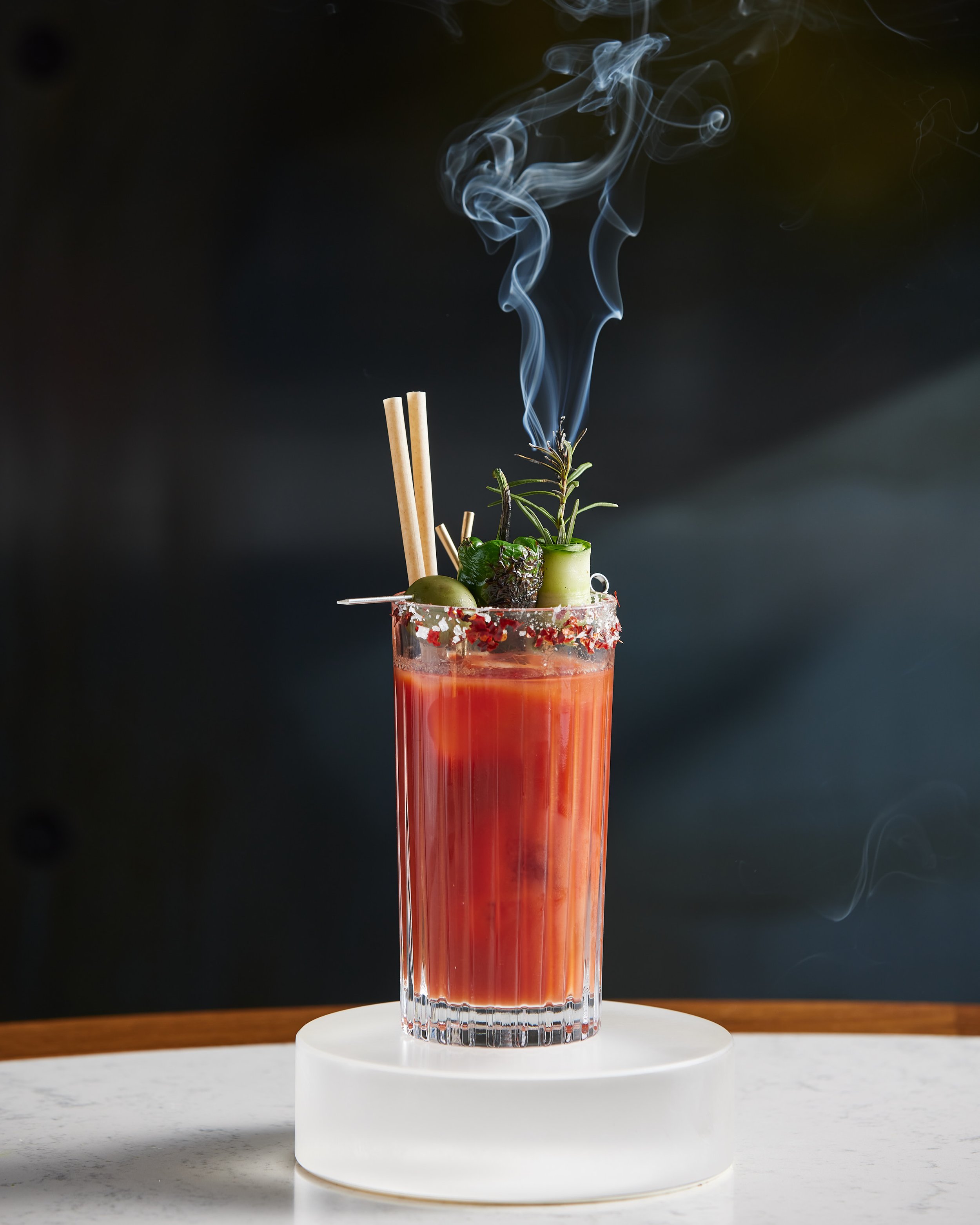 A tall glass of red cocktail garnished with herbs, olives, and skewers, with smoke rising from the top, on a white platform.