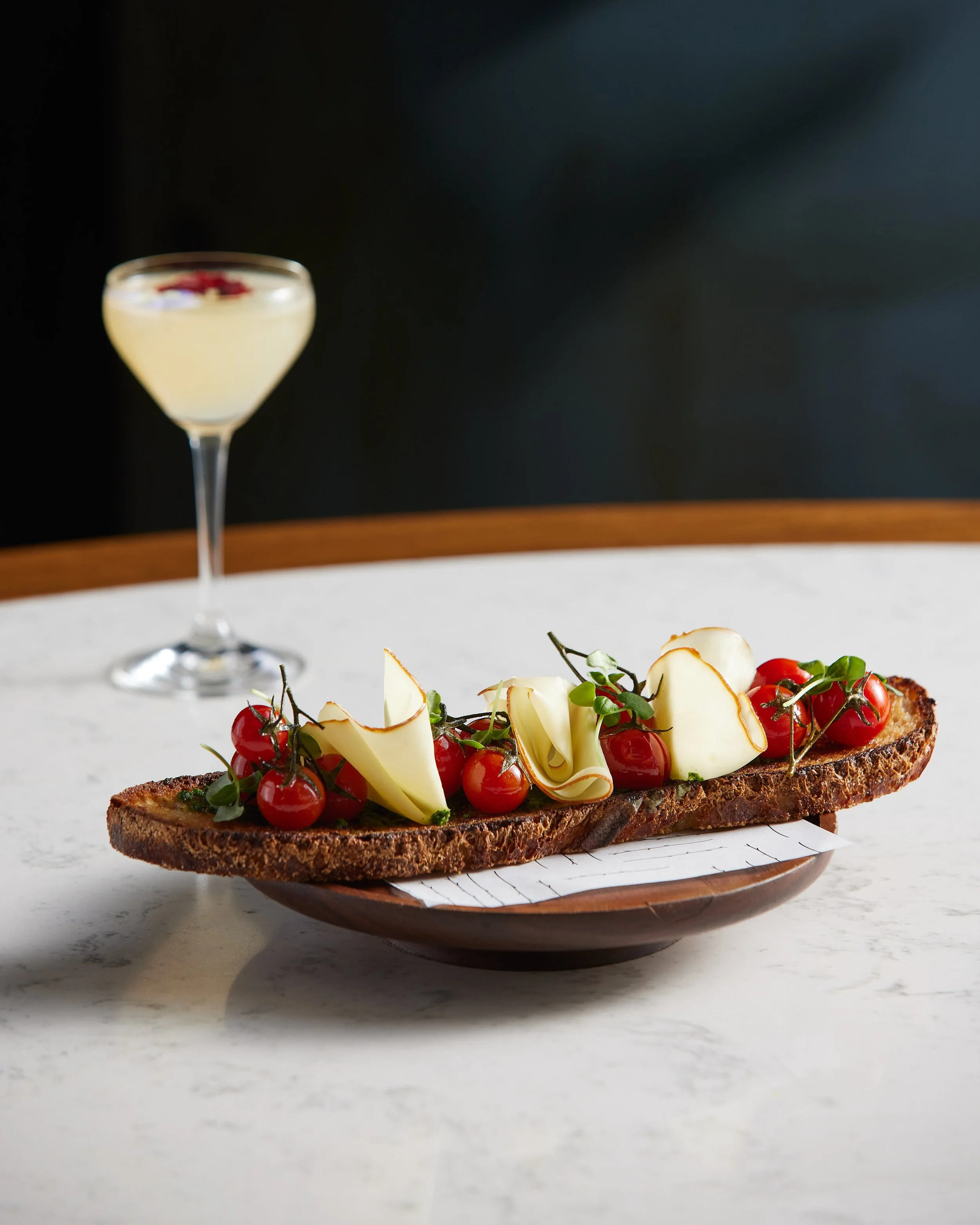 Toast topped with cherry tomatoes, cheese shavings, and herbs, with a cocktail in the background