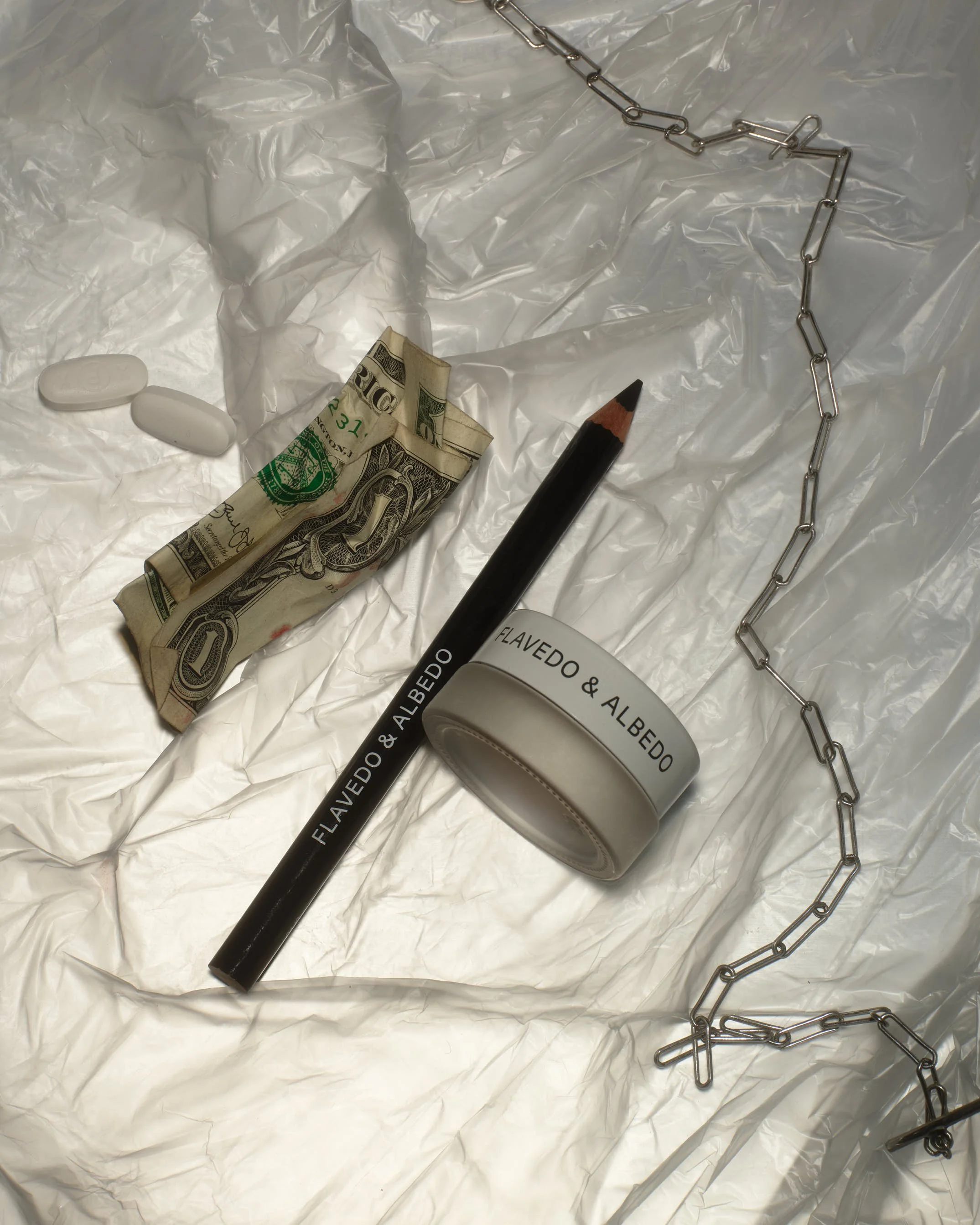 A crumpled piece of white paper with two white pills, a folded ten-dollar bill, a black pen, a white mug with black text, and a metal chain.