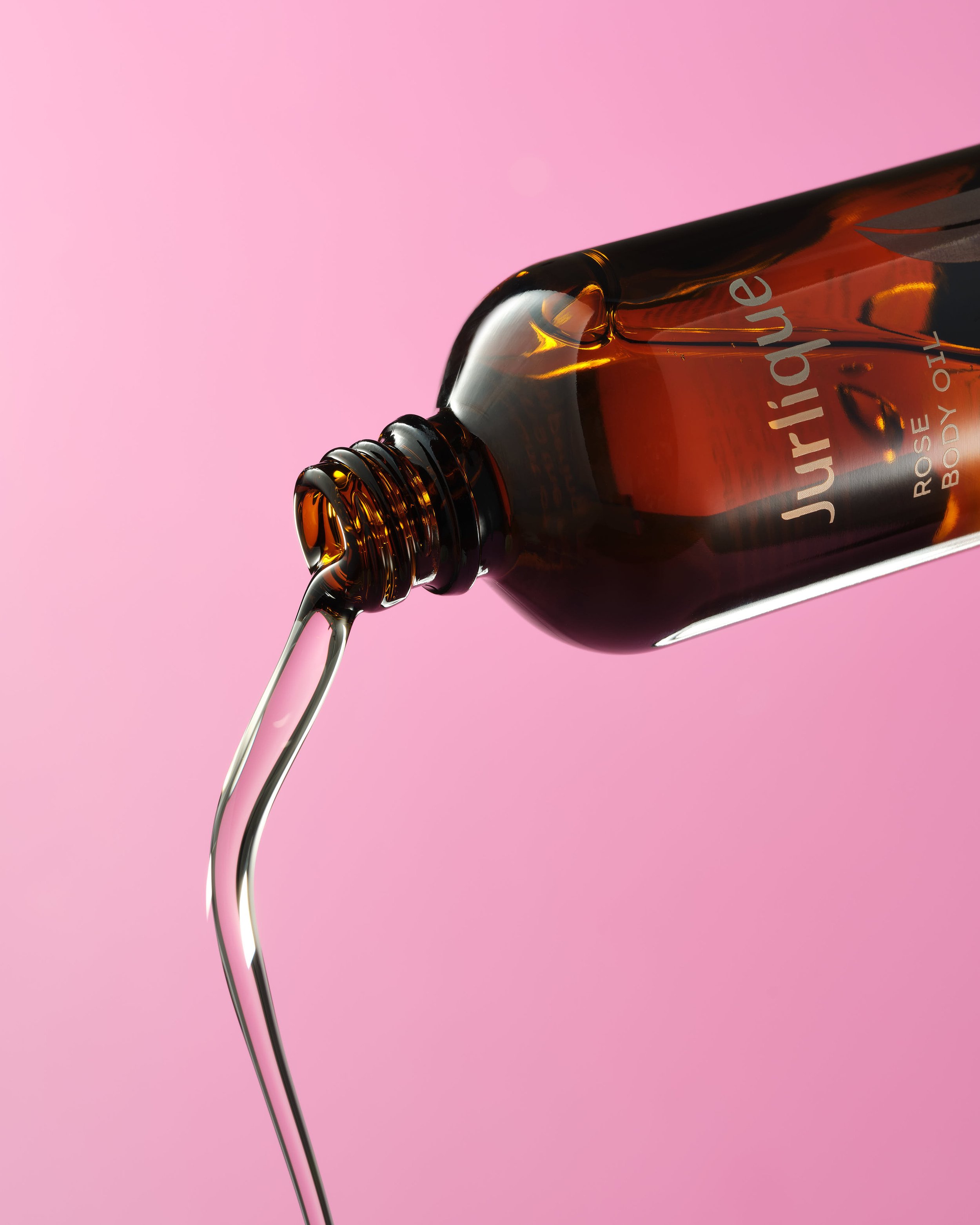 Close-up of a brown glass bottle labeled 'Jurlique' with serum or oil being poured out, against a pink background.