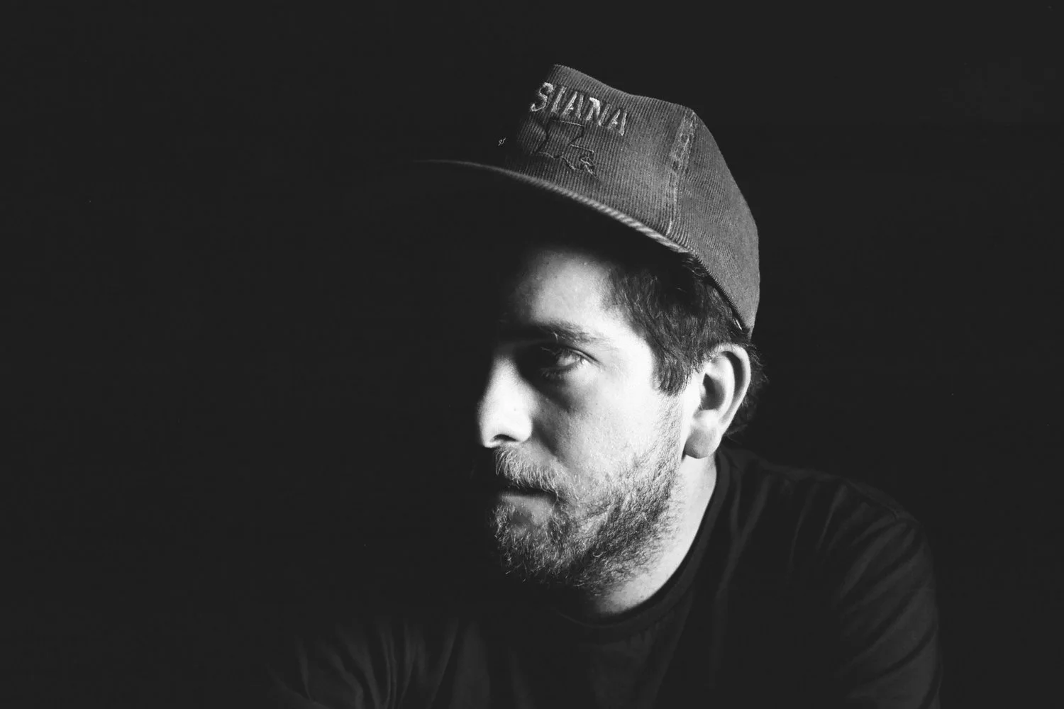 Black and white portrait of a man with a beard wearing a cap, facing left with shadows partially covering his face against a dark background.