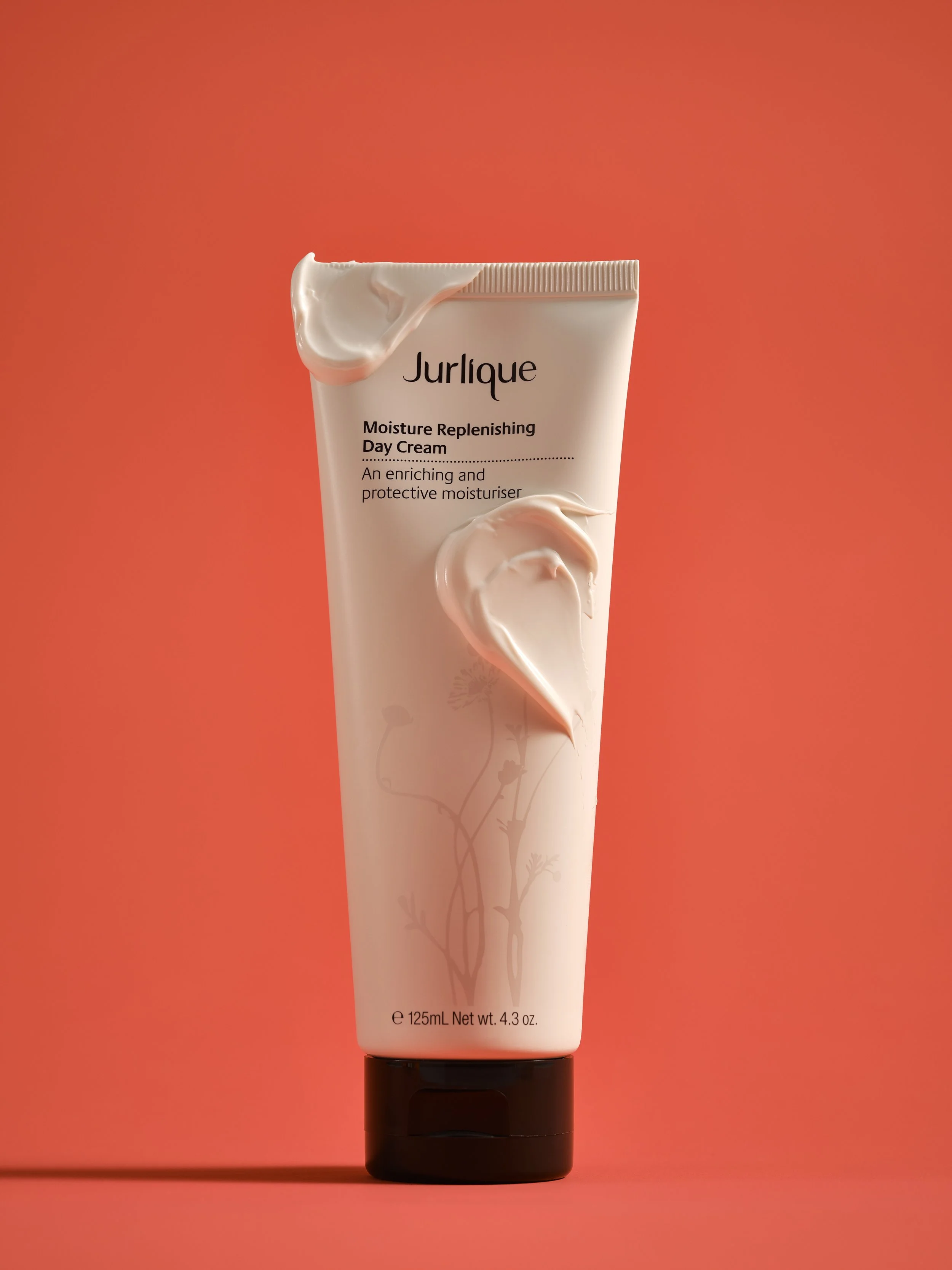 A tube of Jurlique Moisture Replenishing Day Cream with a small amount of cream on the tube's opening, against an orange background.