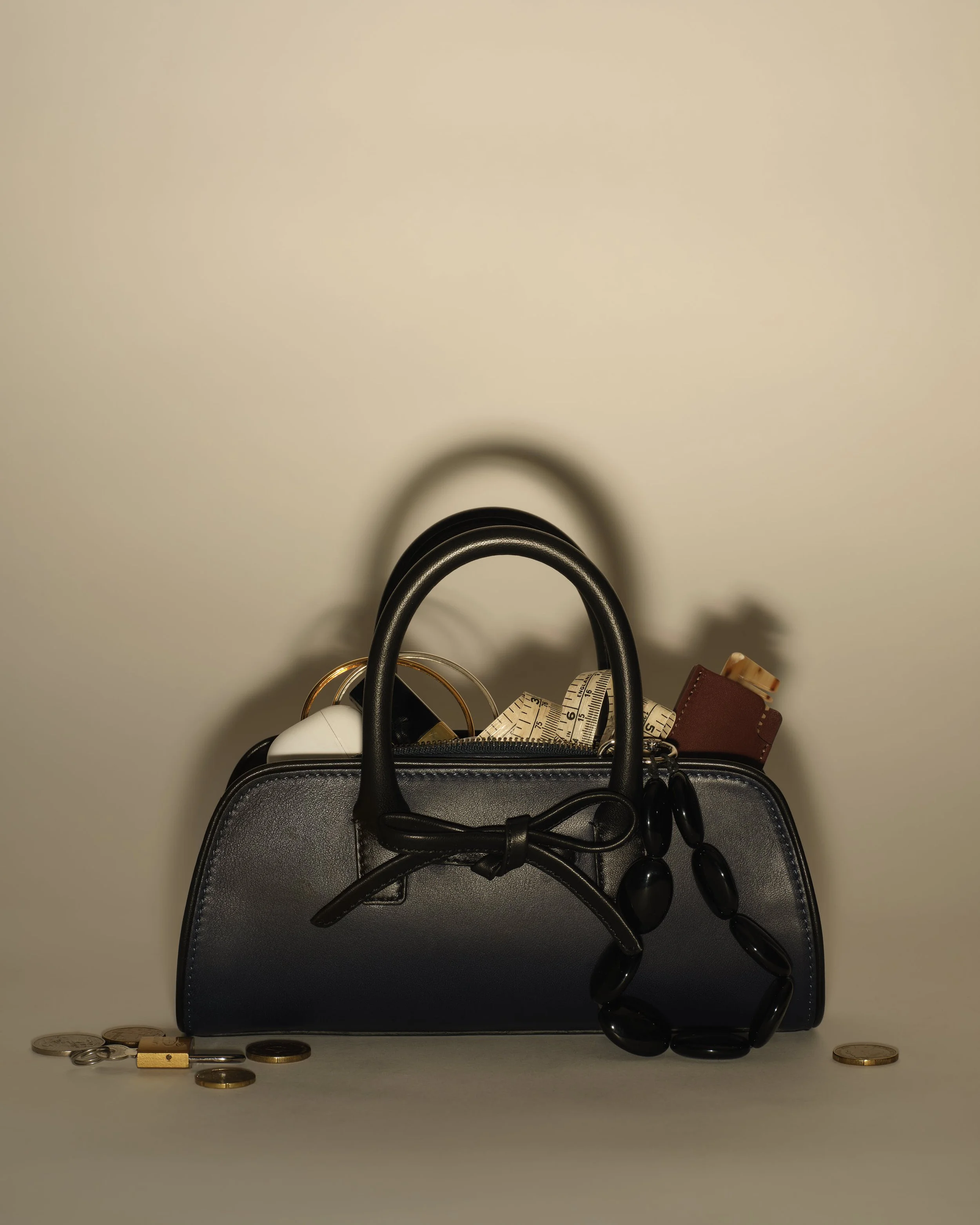 A black purse filled with various objects, including a measuring tape, a brown leather wallet, and a pair of black earbuds, surrounded by scattered coins and keys against a plain beige background.