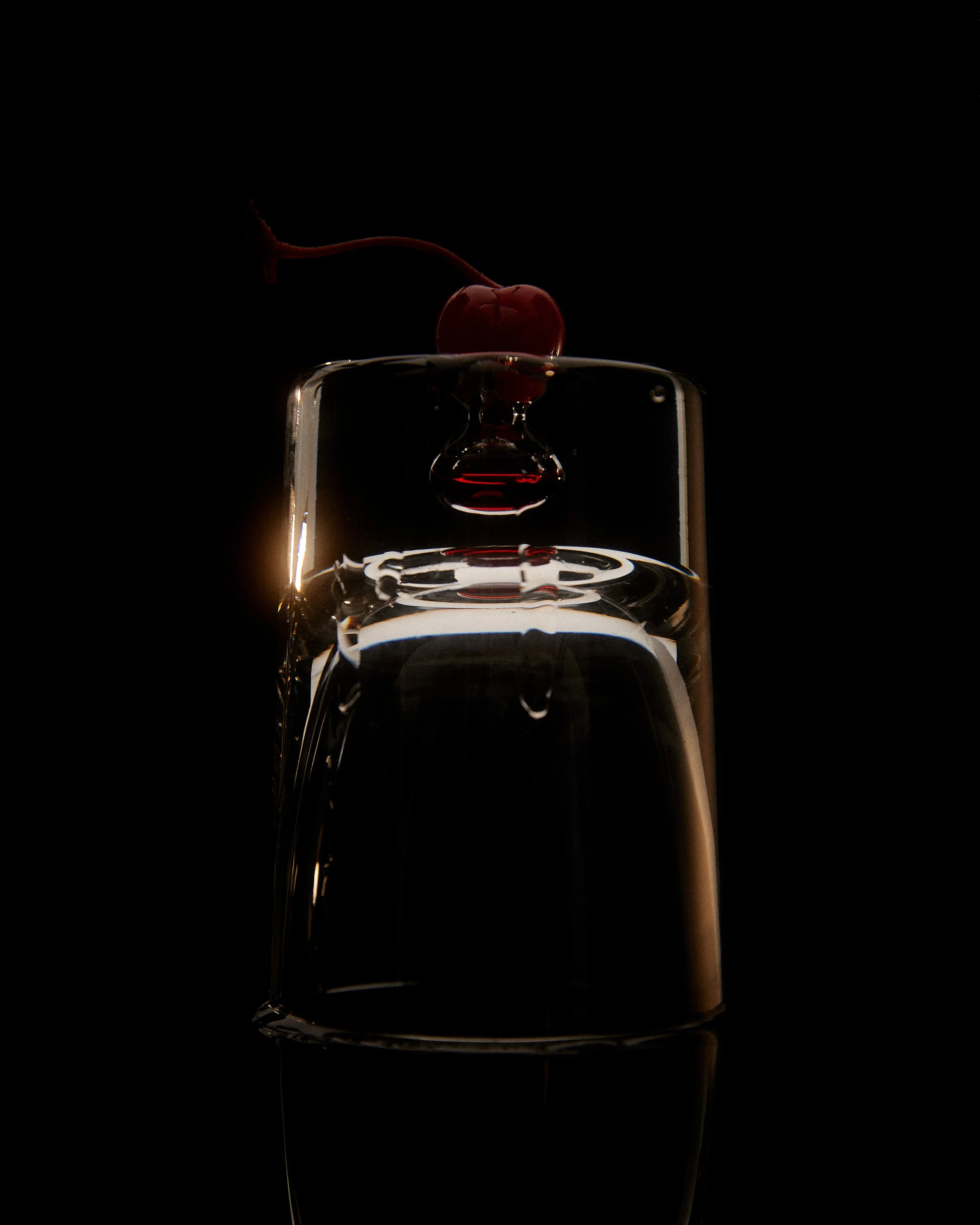 Close-up of a glass of water with a cherry and a drop of water falling from the cherry onto the glass, against a dark background.