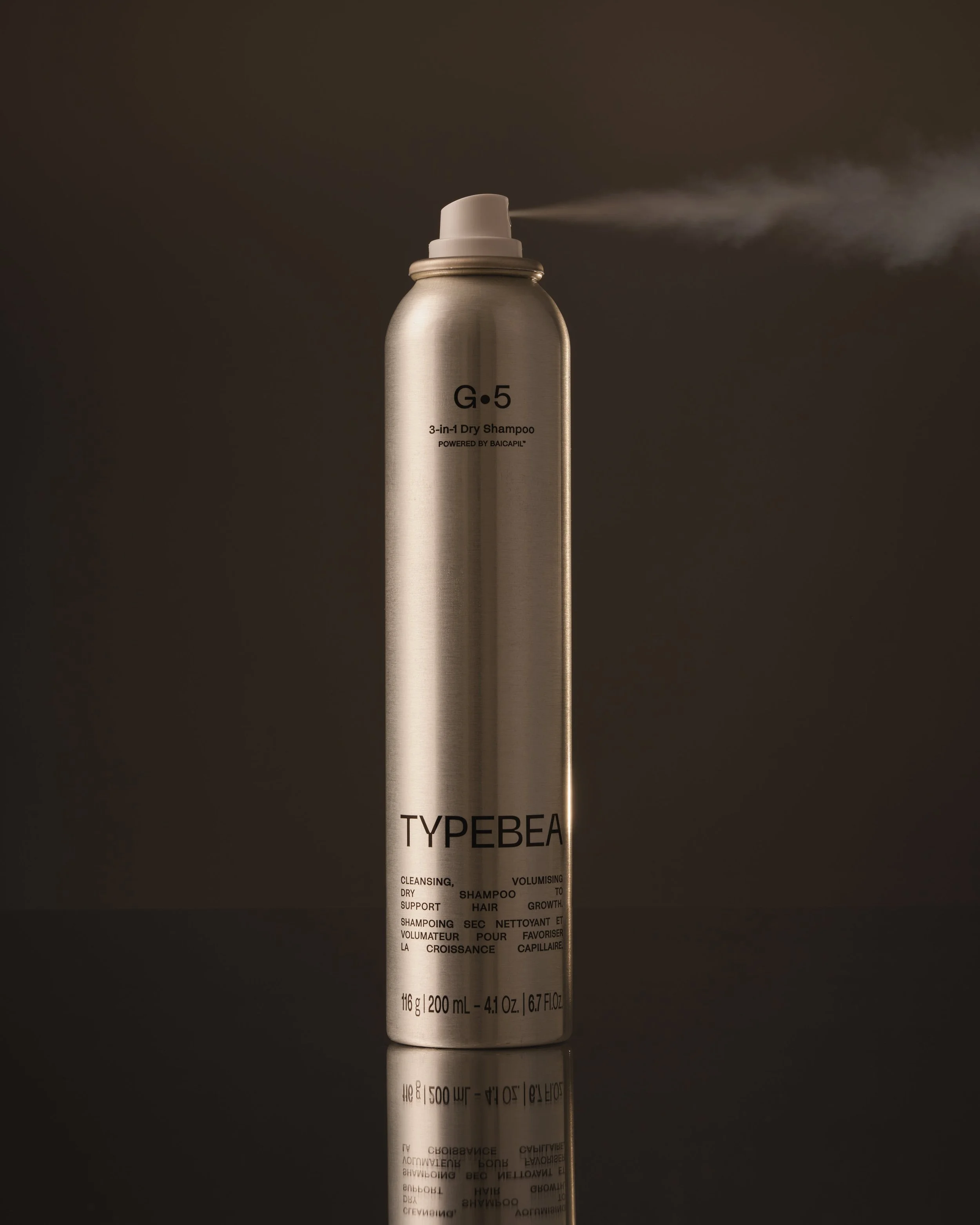 A silver cylindrical can of TYPEBEA 3-in-1 dry shampoo with a white spray nozzle, emitting a spray mist against a dark background.