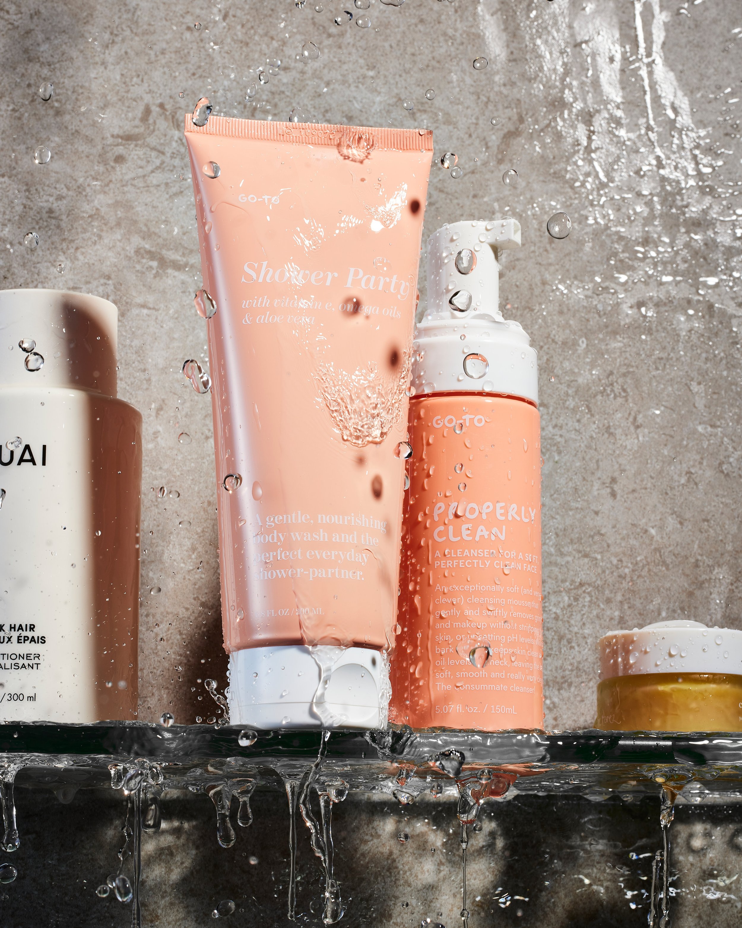 Rain droplets on various skincare and body wash bottles placed on a black shelf against a gray textured background.