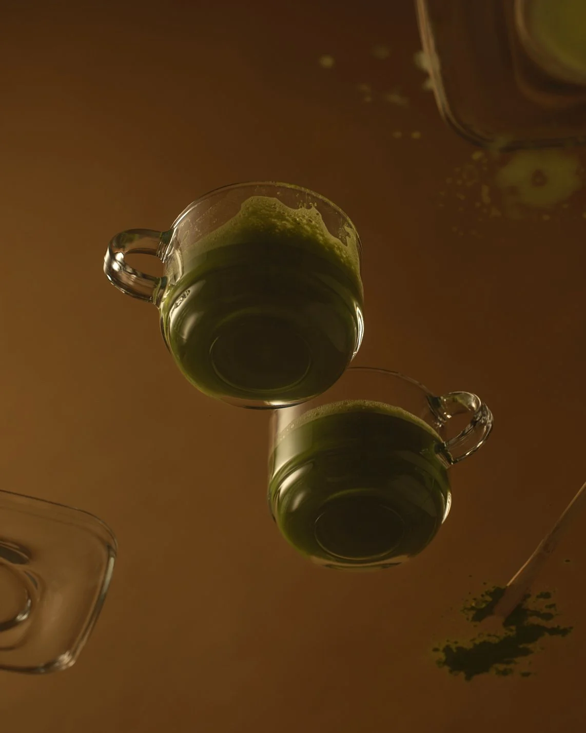 Two glass cups with green liquid and lime slices floating inside, placed on a reflective surface with some scattered green powder or herbs and a straw nearby.
