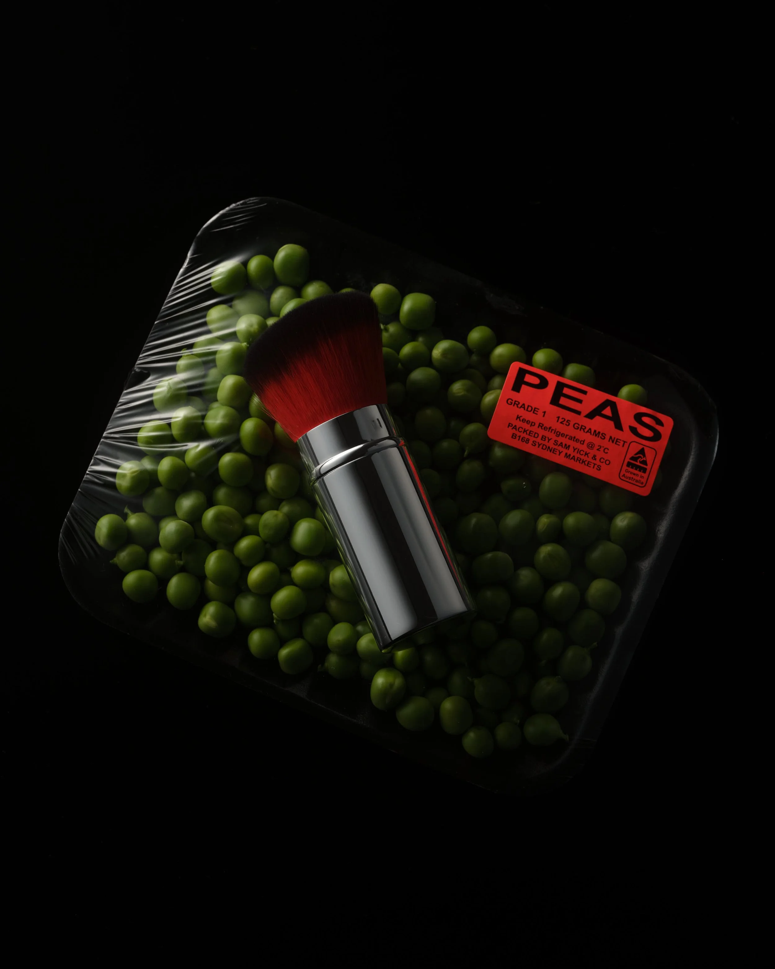 Pack of green peas in a clear plastic bag with a red sticker label, and a metallic makeup brush with red bristles resting on the peas.