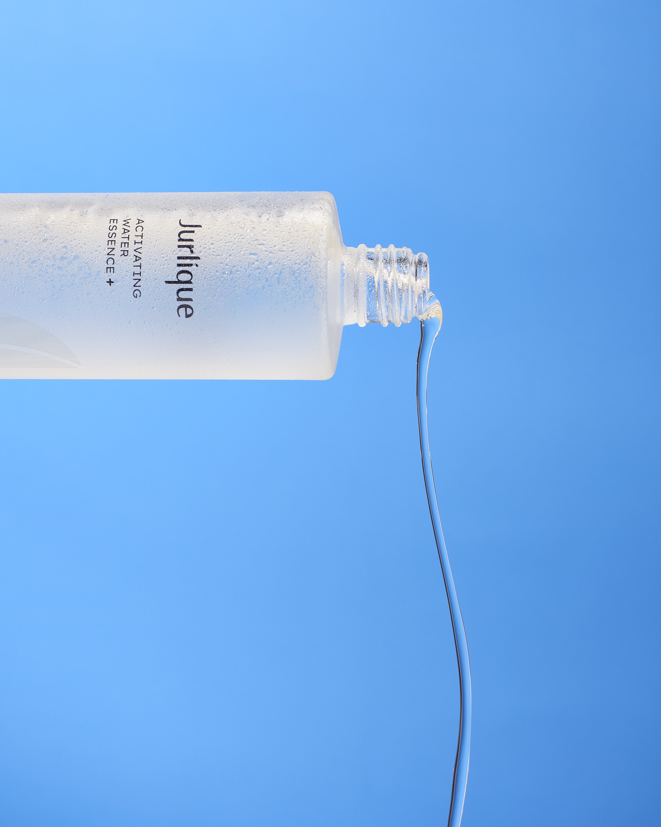 A transparent bottle with water droplets, labeled 'Jurlique Active Water Essence,' tipped to pour a thin stream of liquid against a bright blue background.