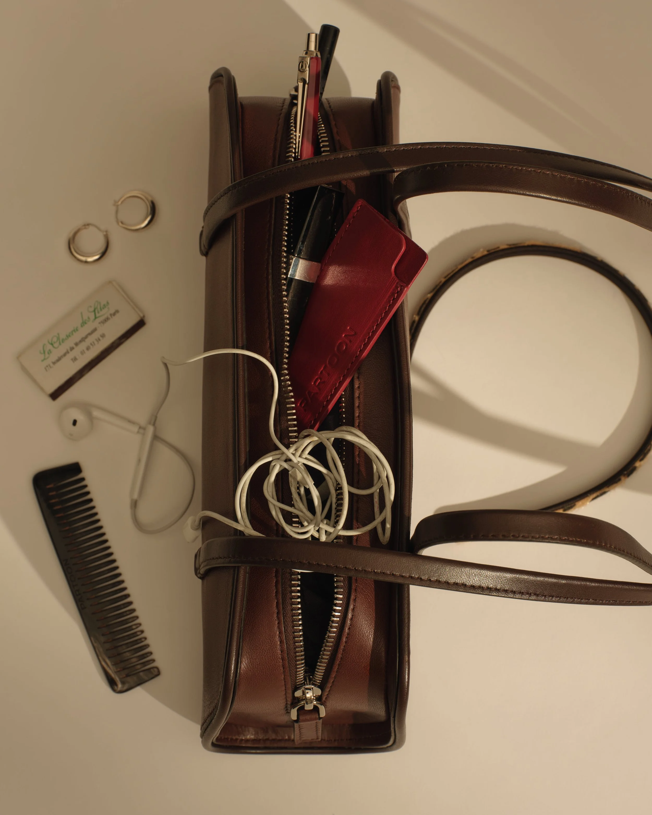 A brown leather handbag with a zipper compartment filled with various items, including a lipstick, a red cardholder, and a pen. The handbag is placed on a white surface with a black comb, silver hoop earrings, a white charging cable, and a small white card nearby.
