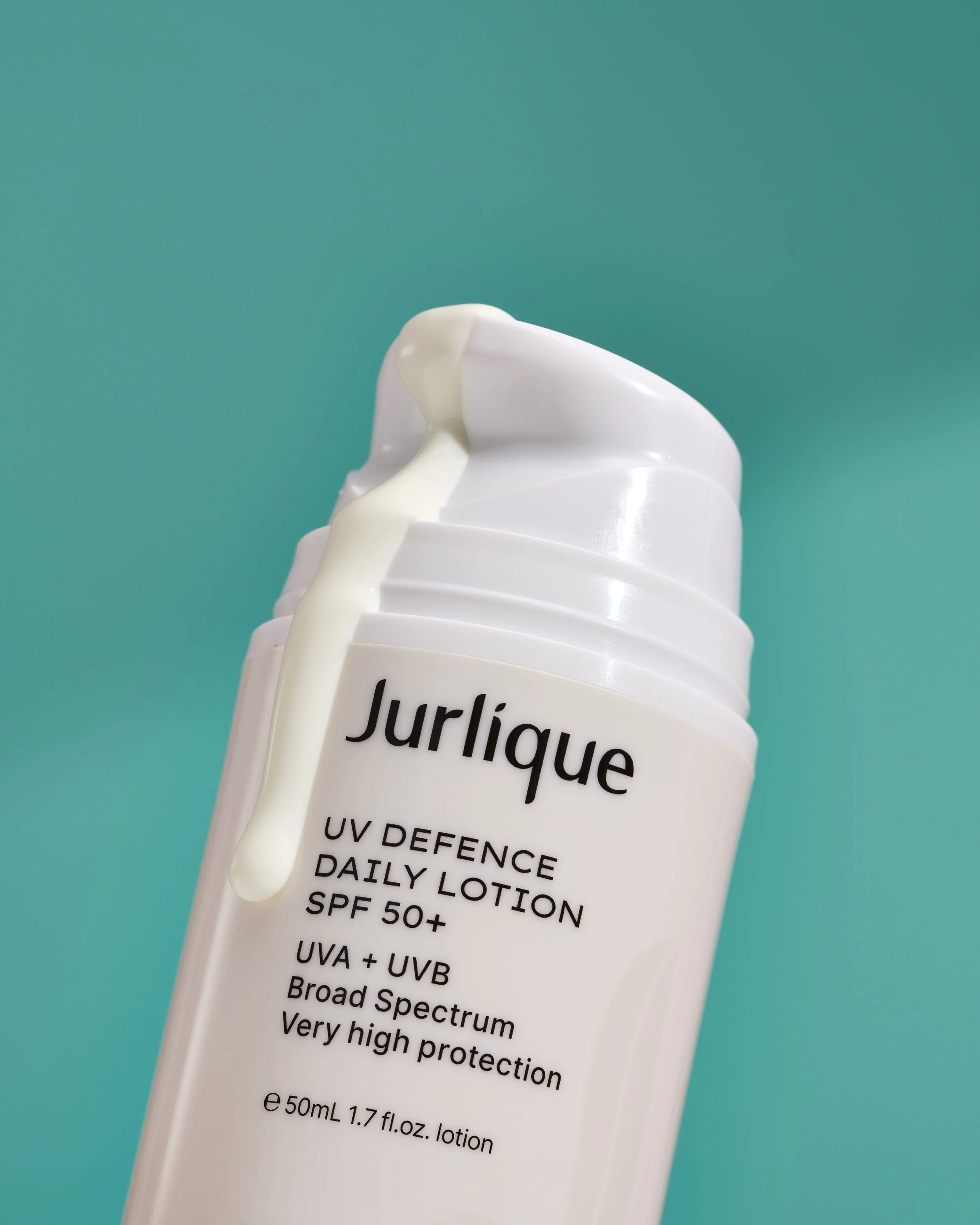 Close-up of a white lotion bottle with the label 'Jurlique' against a teal background. Lotion is spilling over the top of the bottle.