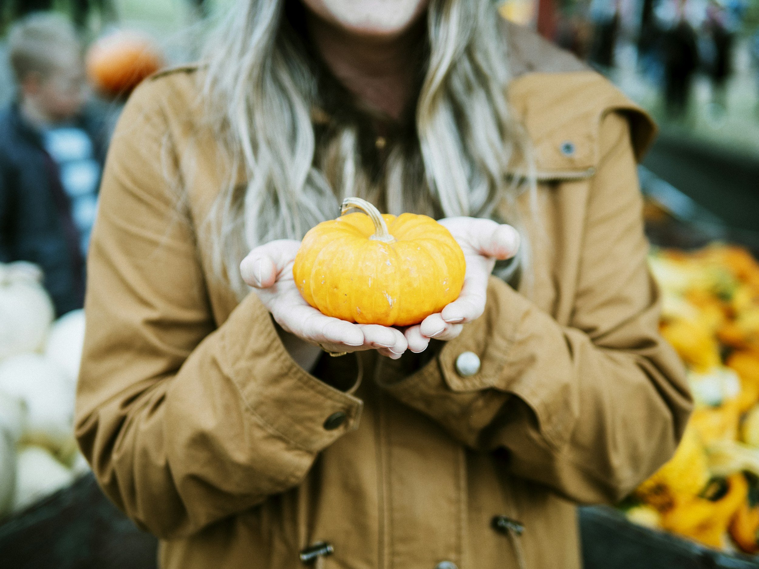 A woman in a brown jacket holding a small orange pumpkin in her hands in a market or grocery store.