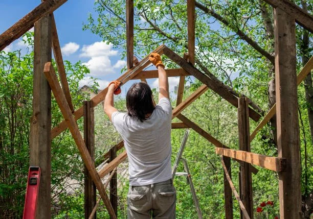 Person building a wooden frame for a structure outdoors with trees and blue sky in the background.