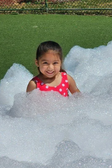A young girl in a red polka dot swimsuit smiling while sitting in a bubble-filled pool.