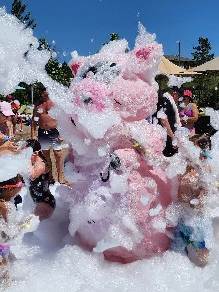 Children and adults at a foam party outdoors, with a person dressed in a pink bunny costume covered in foam.