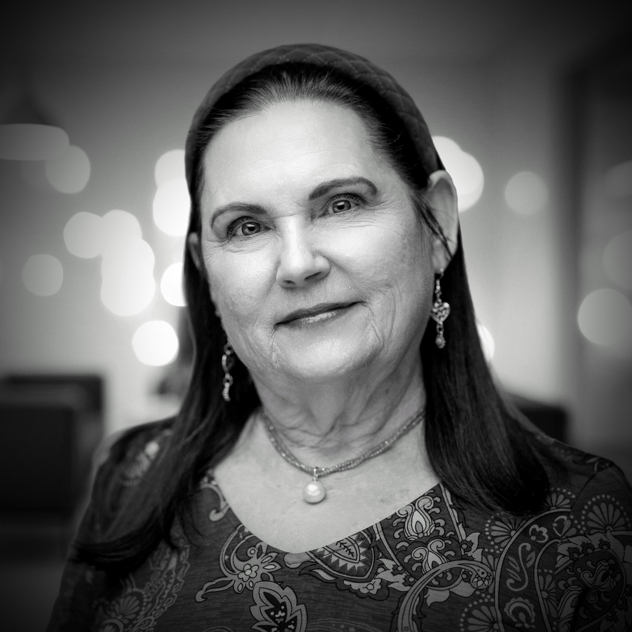 Black and white portrait of an older woman with long dark hair, earrings, and a pearl necklace, smiling softly with a blurred background.