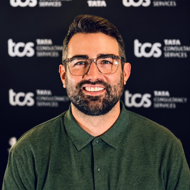 A man with glasses, a beard, and dark hair, wearing a green polo shirt, smiling in front of a backdrop with the logos and text for TCS Tata Consulting Services.